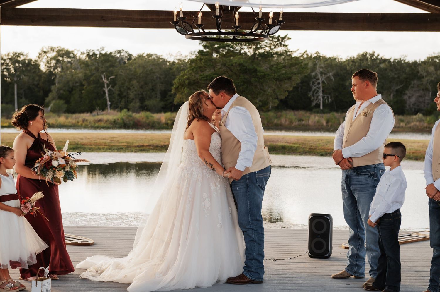 Bride and groom share first kiss at outdoor waterfront wedding ceremony with wedding party in burgundy and tan attire.