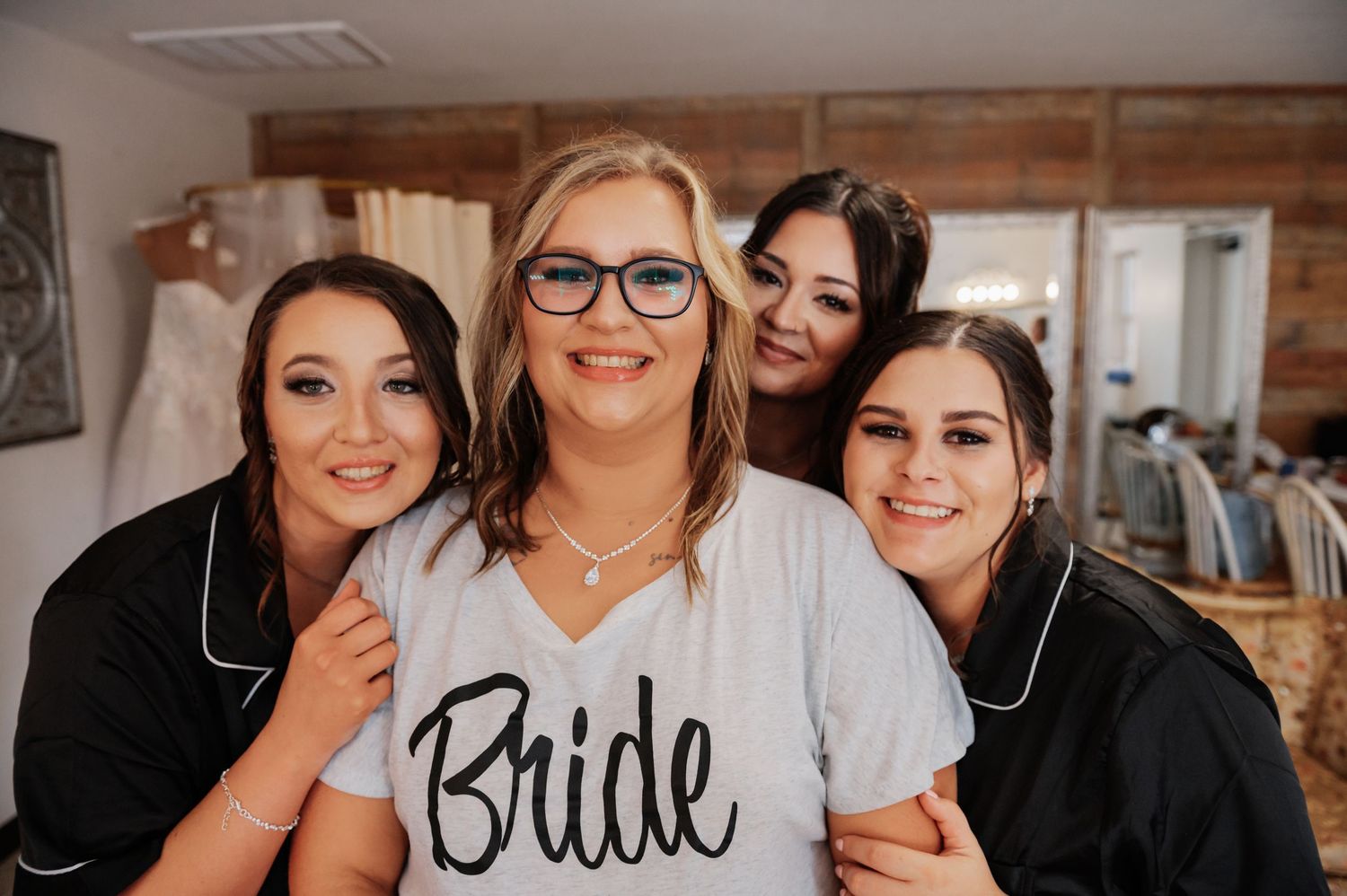 Bride in white shirt with bridesmaids in black robes smiling together during wedding preparations.