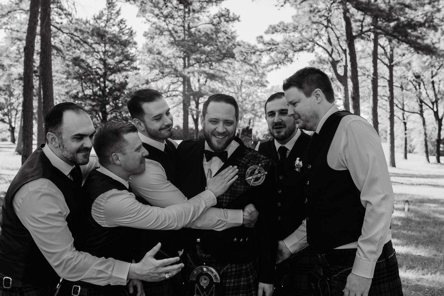Black and white photo of a groom in a kilt celebrating with groomsmen outdoors among pine trees.