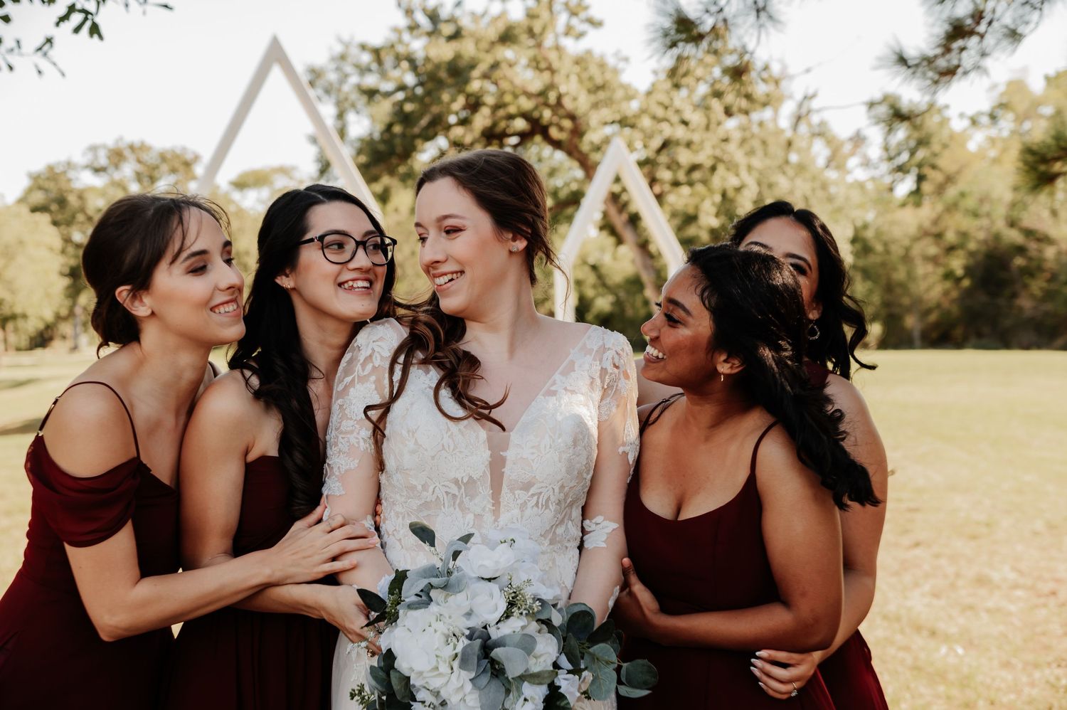 Bride in lace gown holds white bouquet surrounded by bridesmaids in burgundy dresses outdoors at wedding.