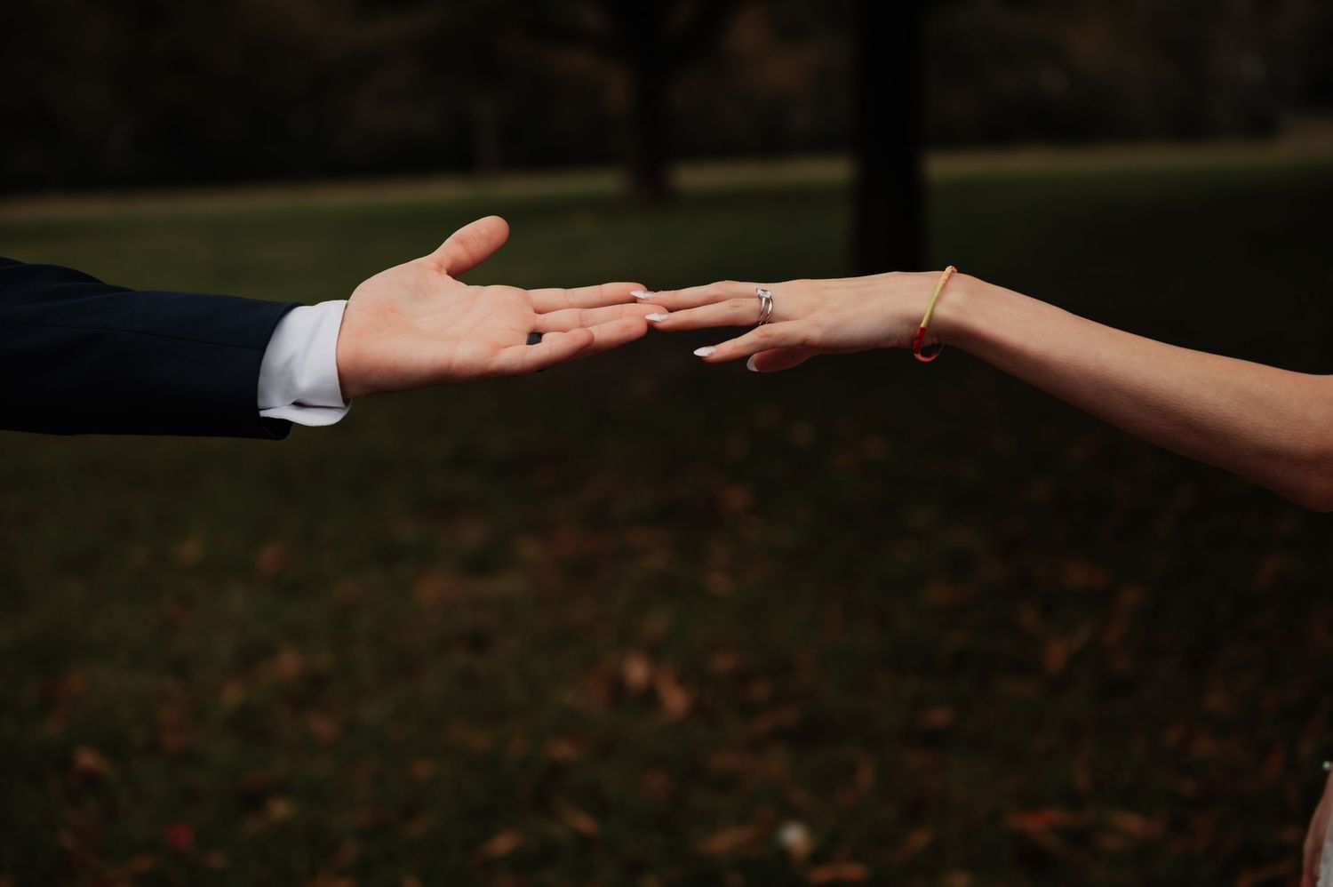 Two hands reaching toward each other outdoors, woman wearing engagement ring, romantic couple moment in autumn park.