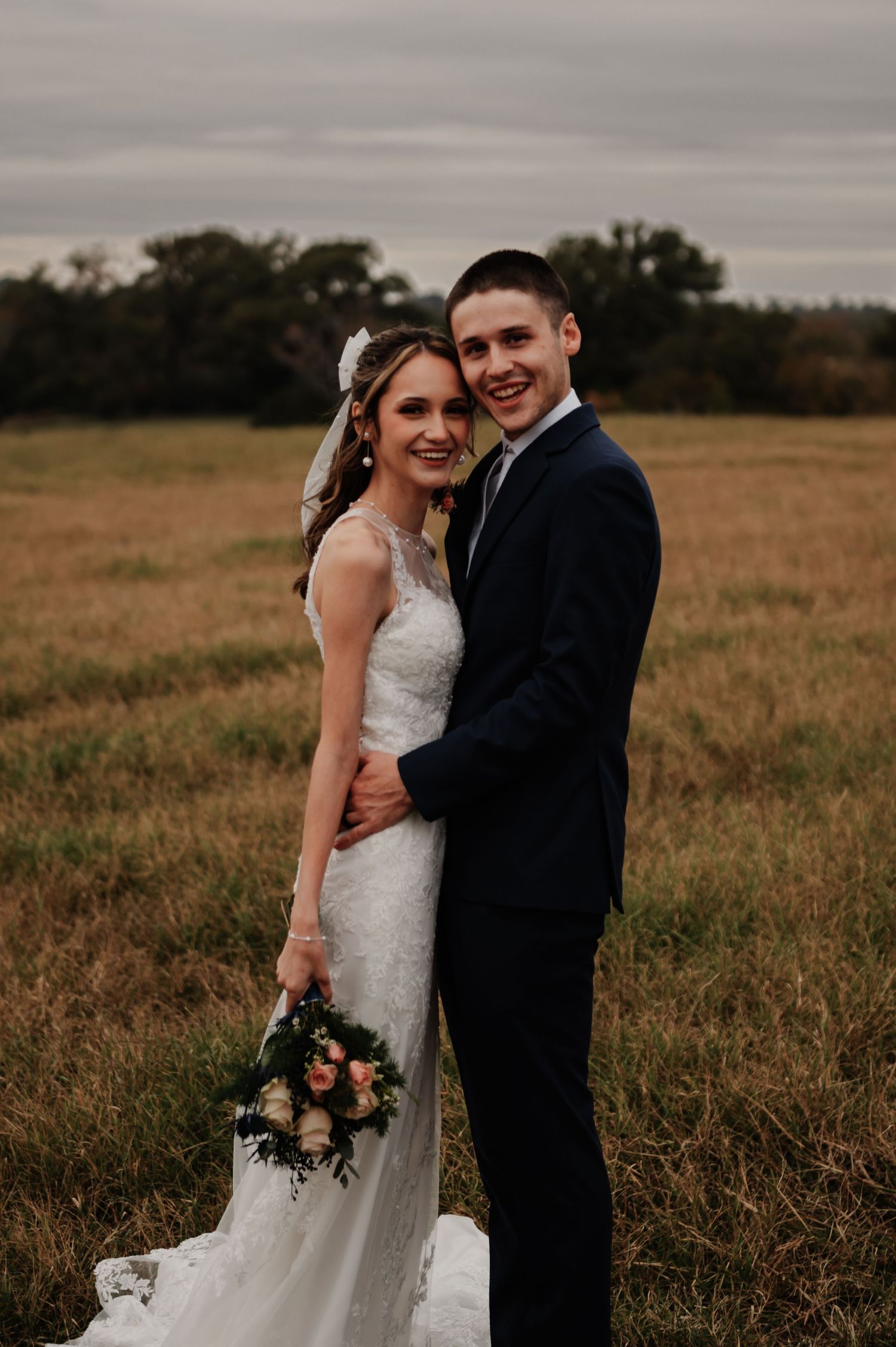 A smiling bride and groom pose in a golden field, she in lace gown with bouquet, he in navy suit.