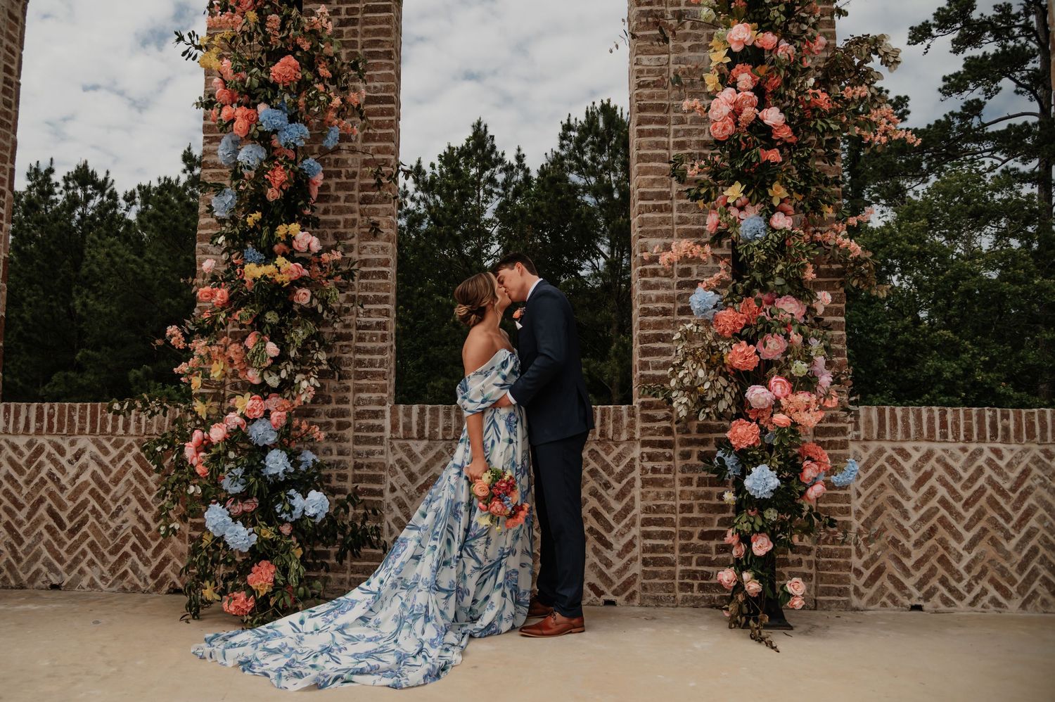 Bride in blue floral gown and groom in navy suit share a kiss before brick columns adorned with colorful flowers.