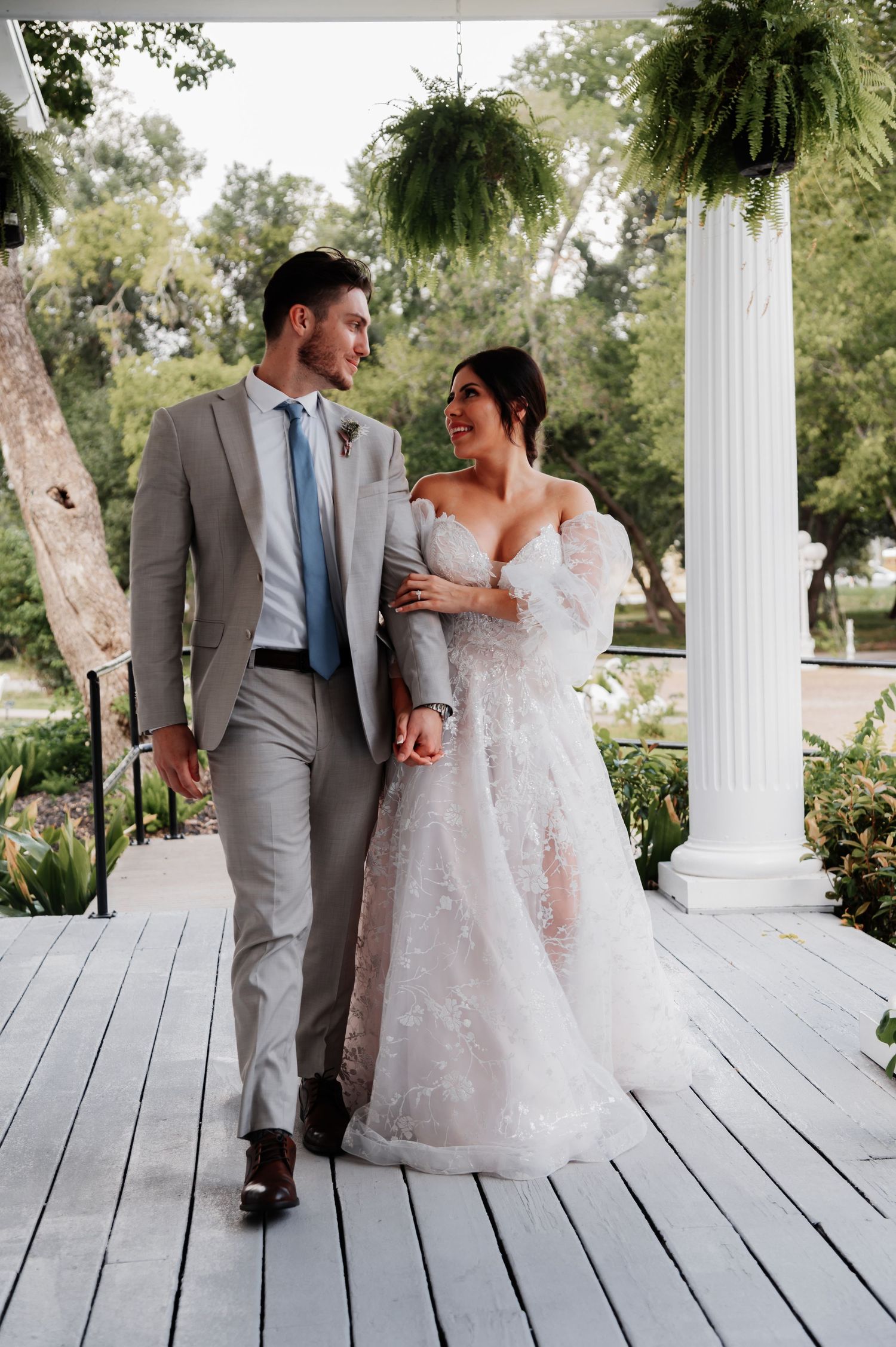 A bride and groom walk hand-in-hand on a white porch, smiling at each other in elegant wedding attire.
