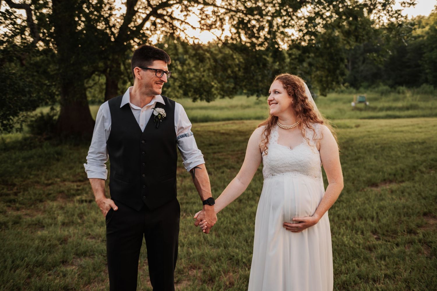 Pregnant bride in white gown holds hands with groom in vest outdoors at golden hour.