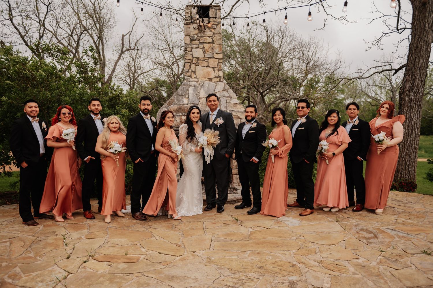Wedding party in coral bridesmaid dresses and black suits posing outdoors on stone patio with rustic stone tower backdrop.