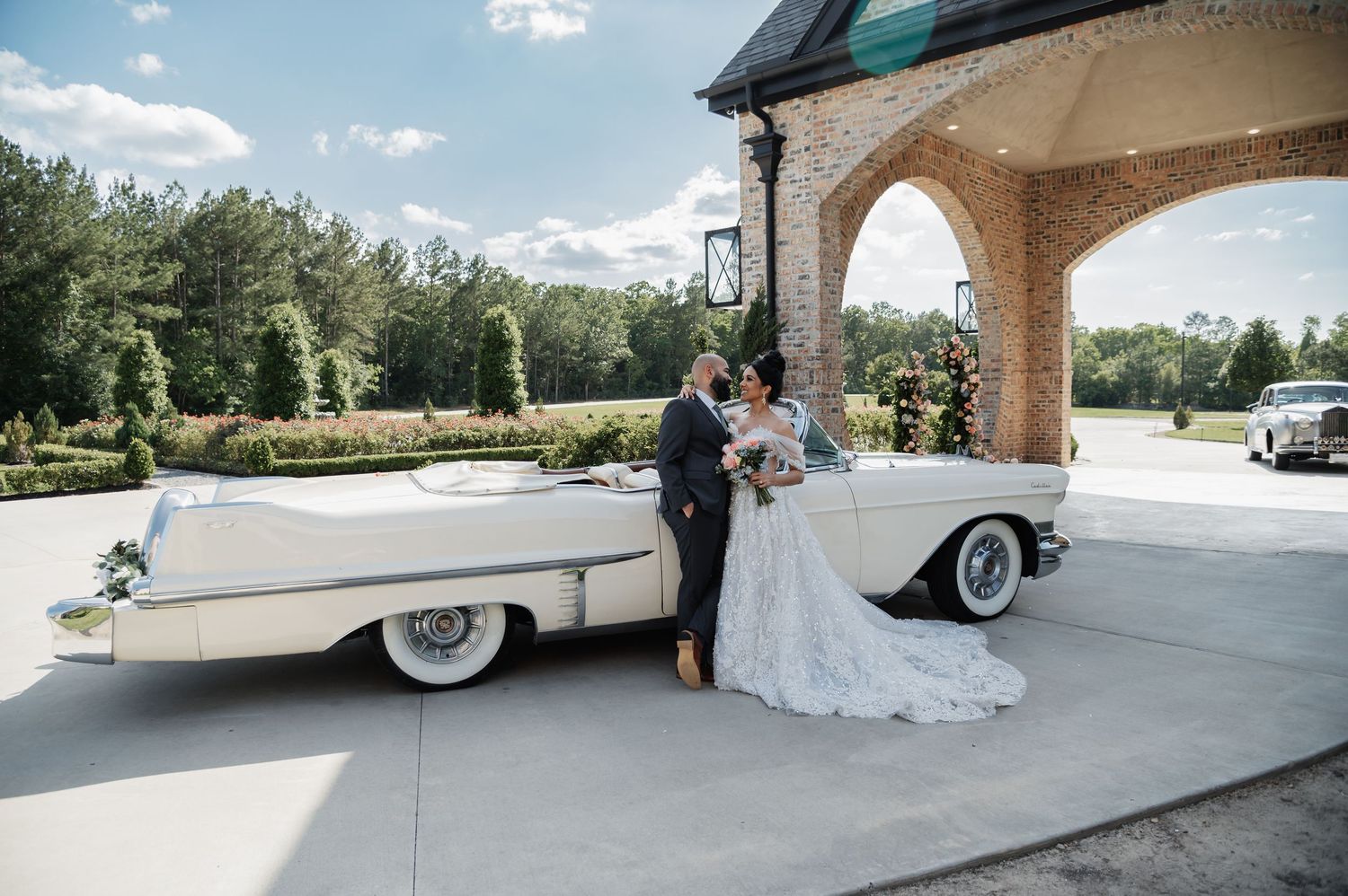 Bride and groom posing beside a classic white vintage convertible car outside an elegant brick wedding venue.