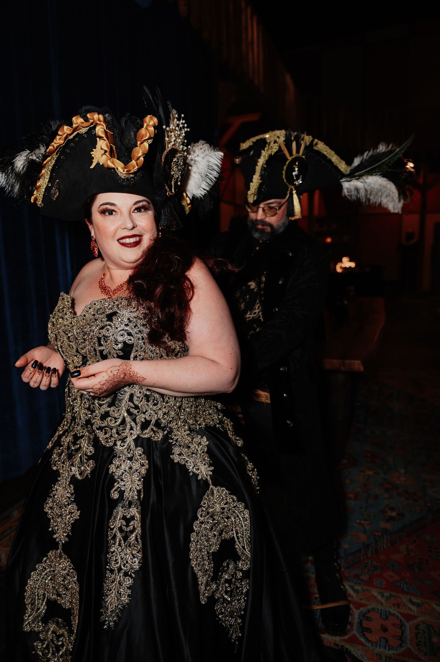 Couple in elaborate black and gold pirate costumes with feathered tricorn hats at a themed wedding event.