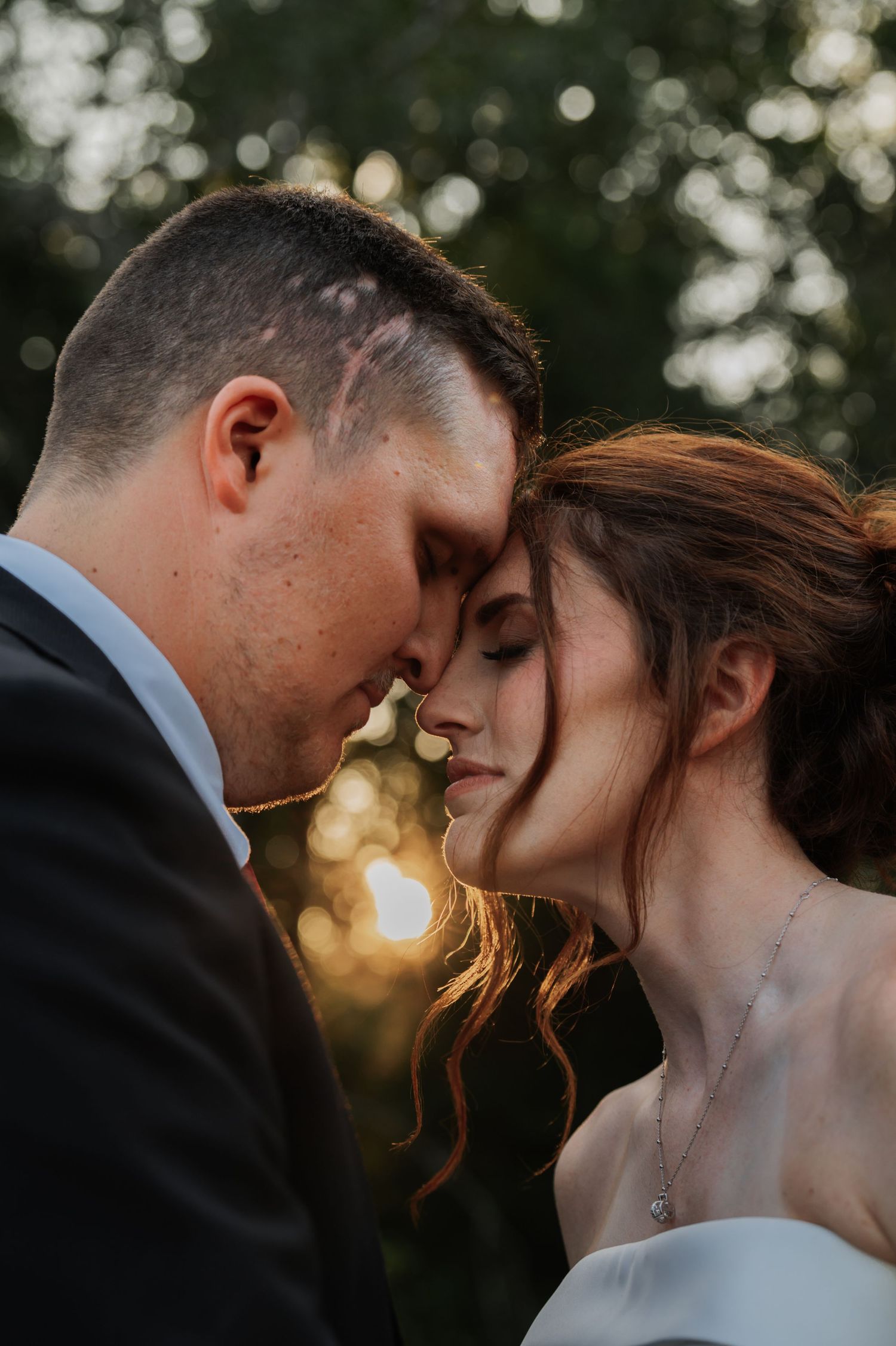 Romantic wedding couple touching foreheads at golden hour sunset, bride in white gown with necklace, groom in dark suit.