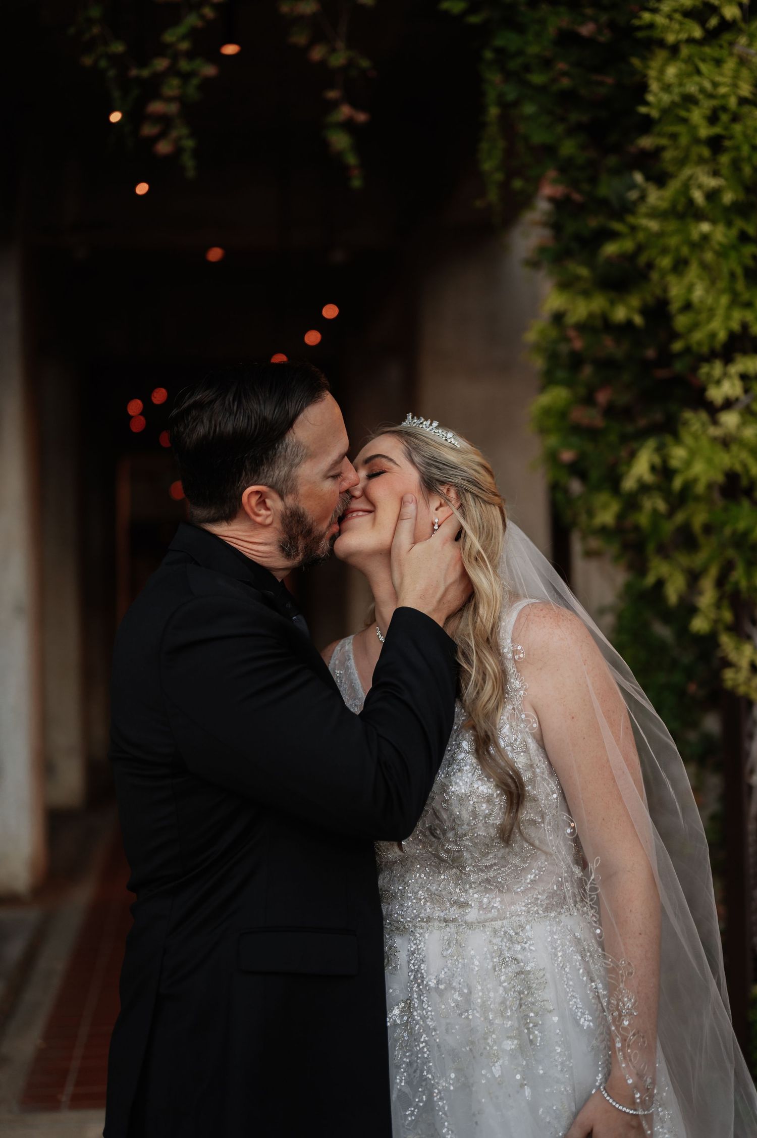 Bride and groom share a romantic kiss outdoors, bride wearing a beaded gown, veil, and tiara.