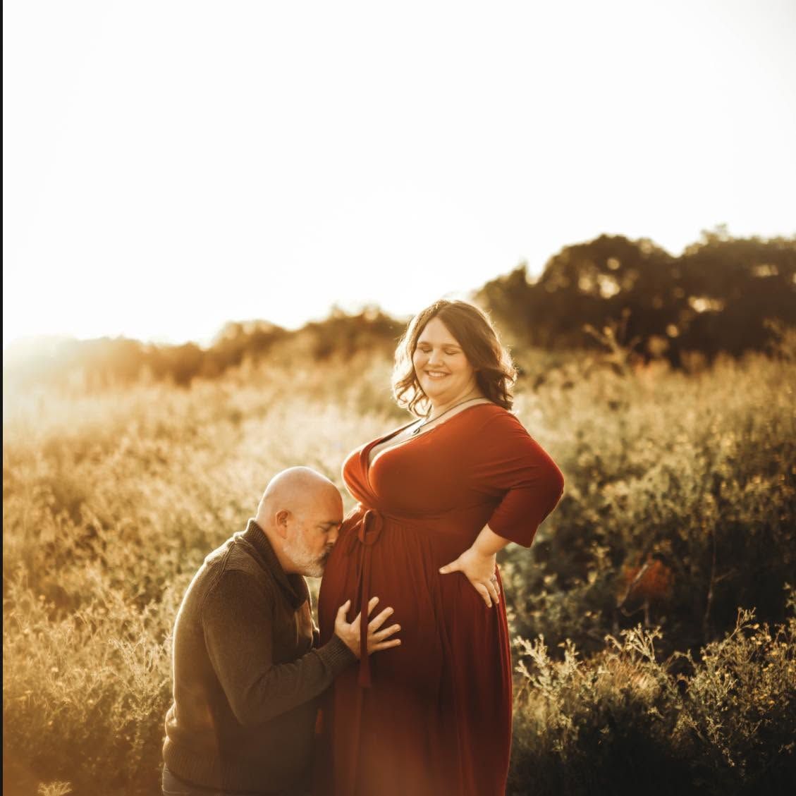 A couple poses in a golden sunlit field, the man tenderly kissing a pregnant woman's belly during a maternity photoshoot.