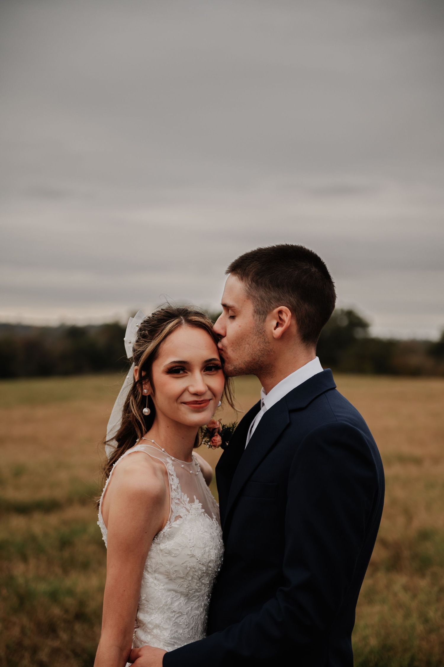 Groom in navy suit kissing bride in lace dress on cheek in golden field on overcast day.