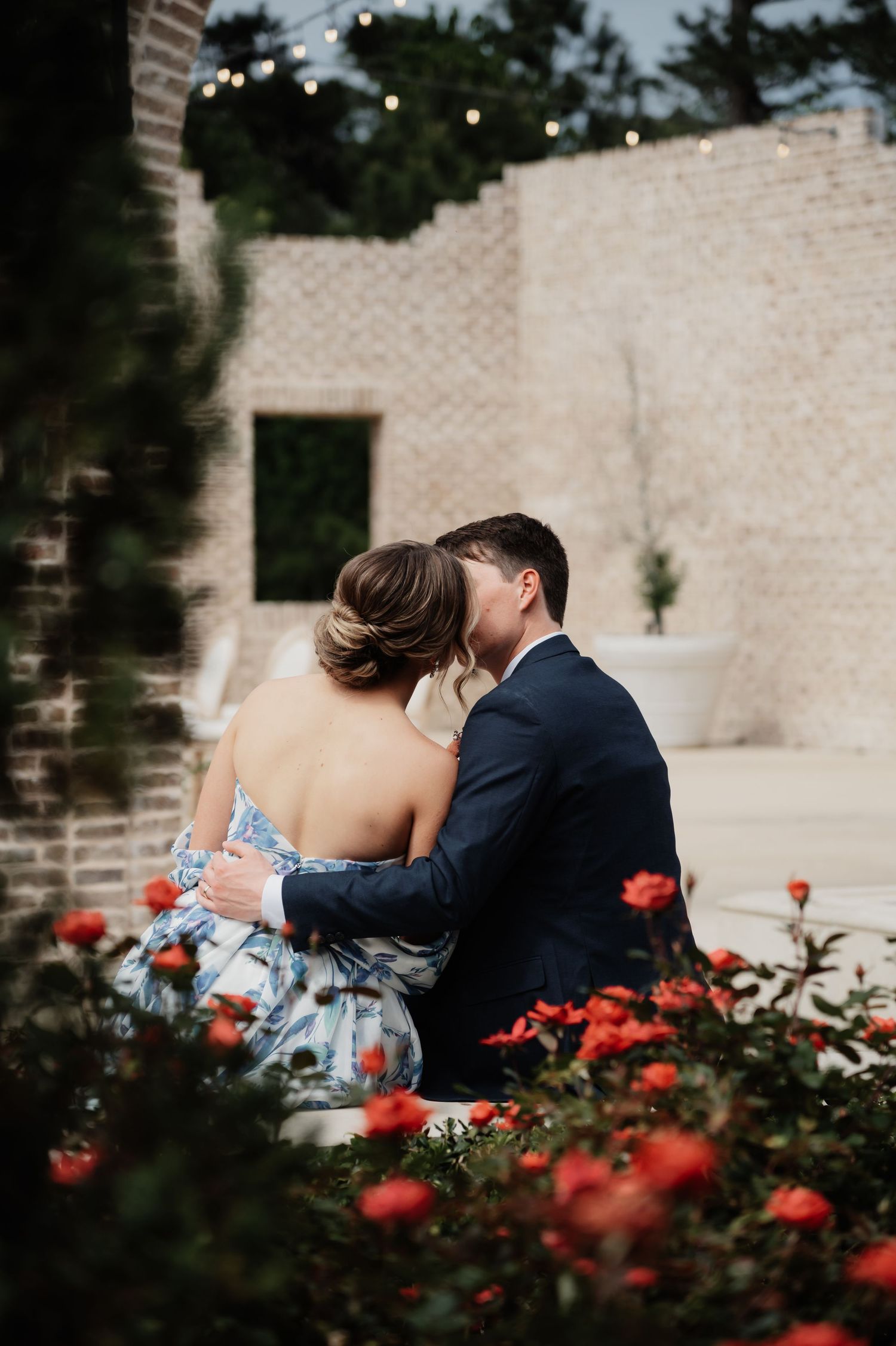 Couple sharing a romantic kiss among red roses in a garden, man in navy suit embracing woman in floral dress.