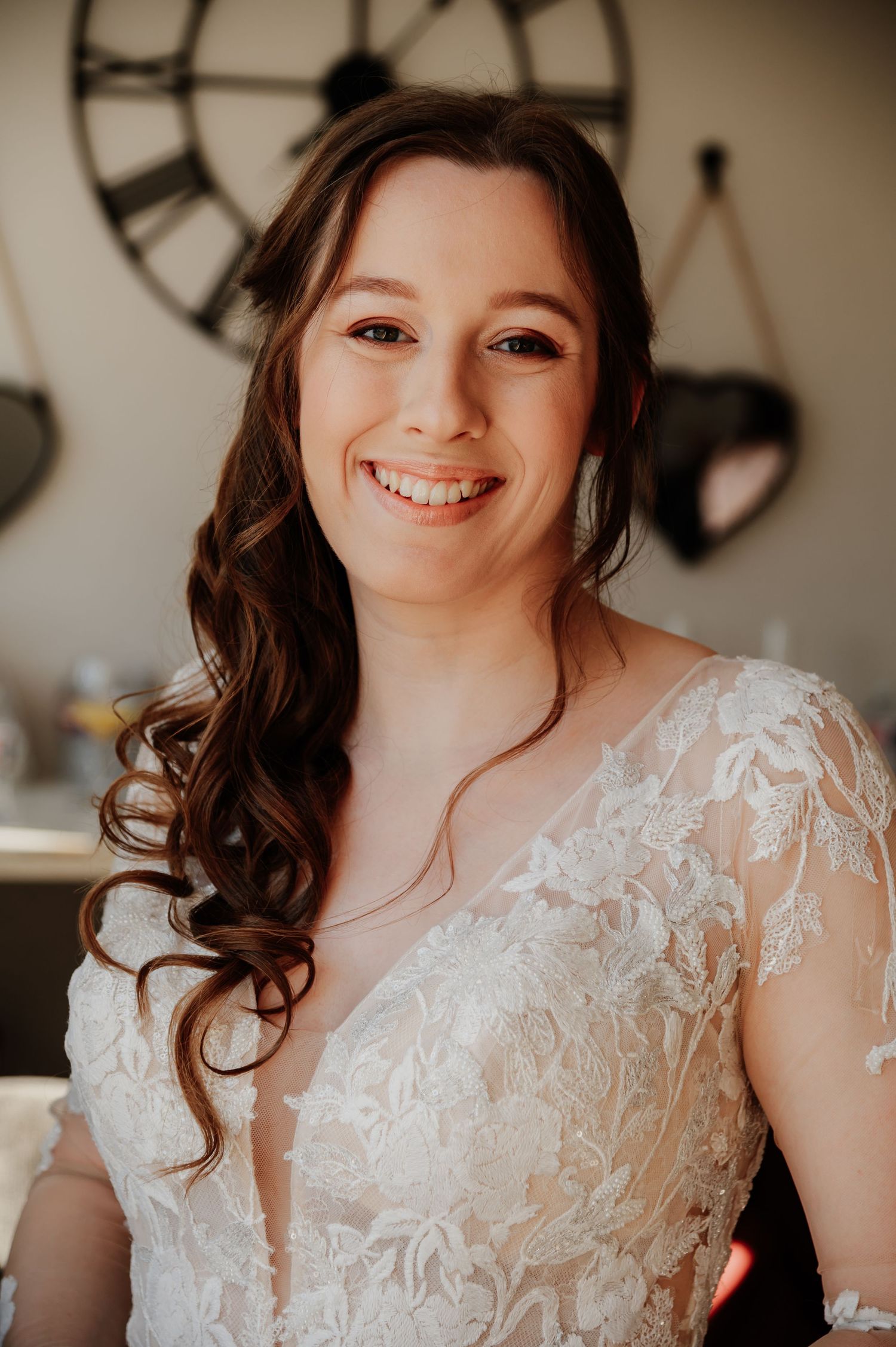Smiling bride in lace wedding dress with curled hair poses indoors near a decorative clock.