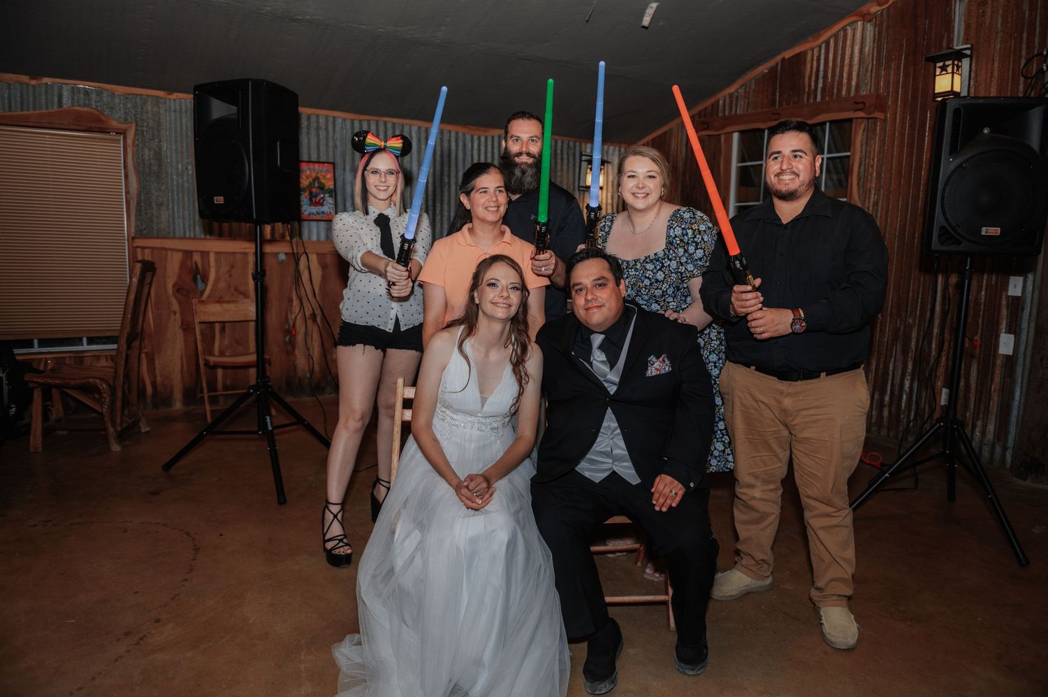 Wedding party poses with colorful lightsabers in a rustic venue, bride and groom centered among Star Wars-themed guests.