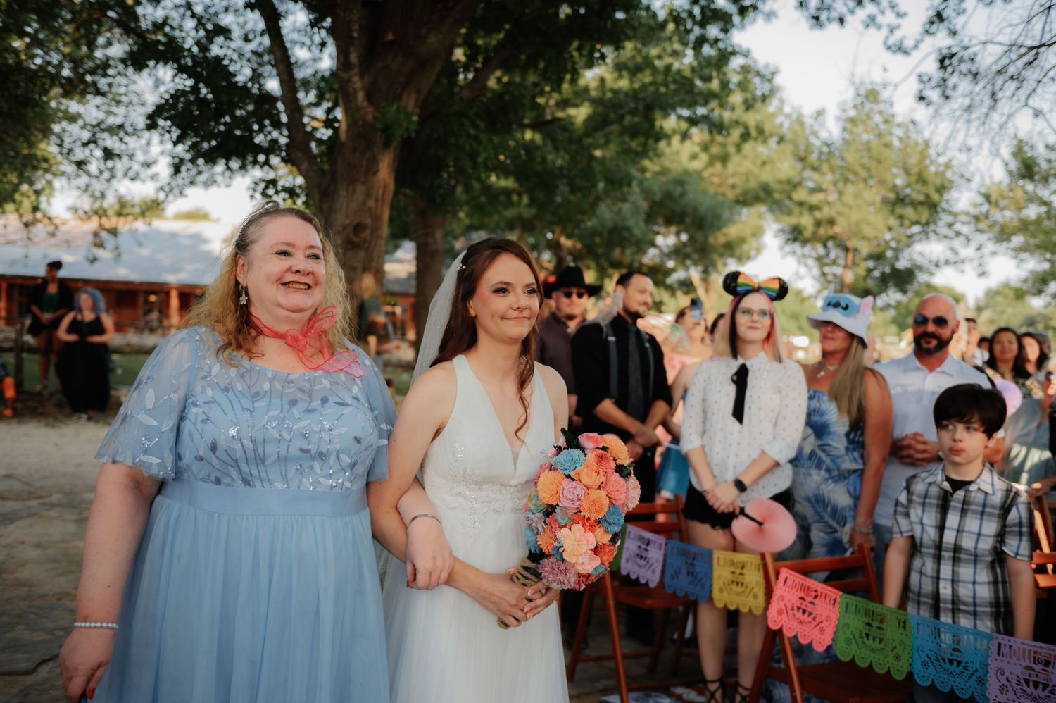Bride in white gown walks aisle with woman in blue dress at outdoor wedding with colorful papel picado decorations.