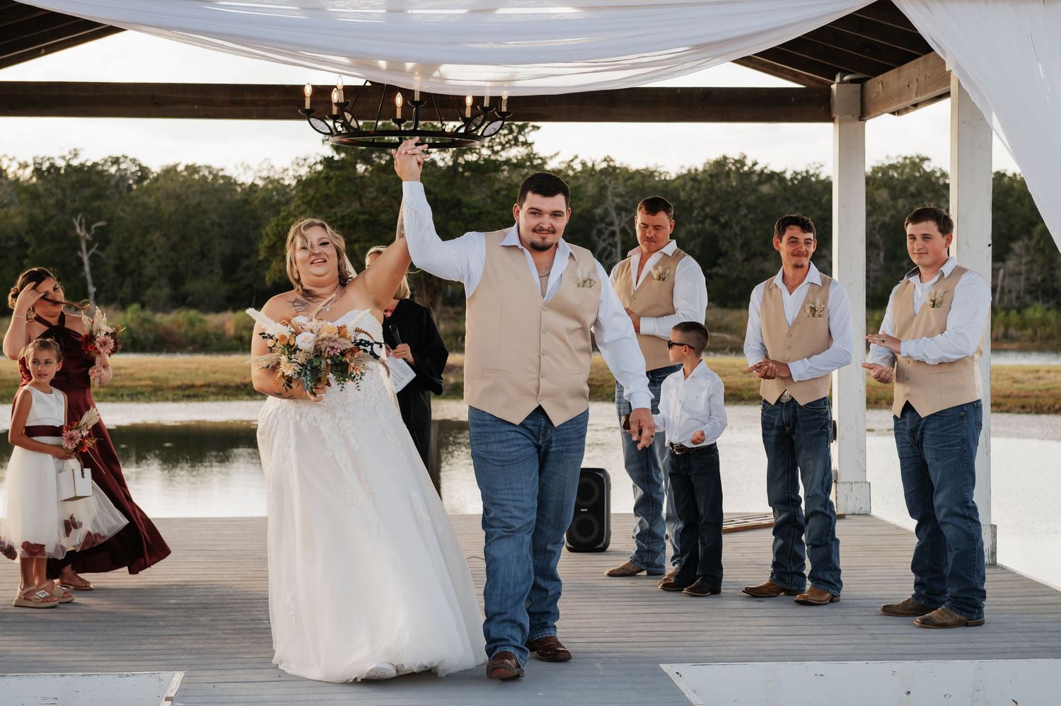 Bride and groom celebrate at outdoor wedding ceremony on dock, surrounded by wedding party in rustic country attire.