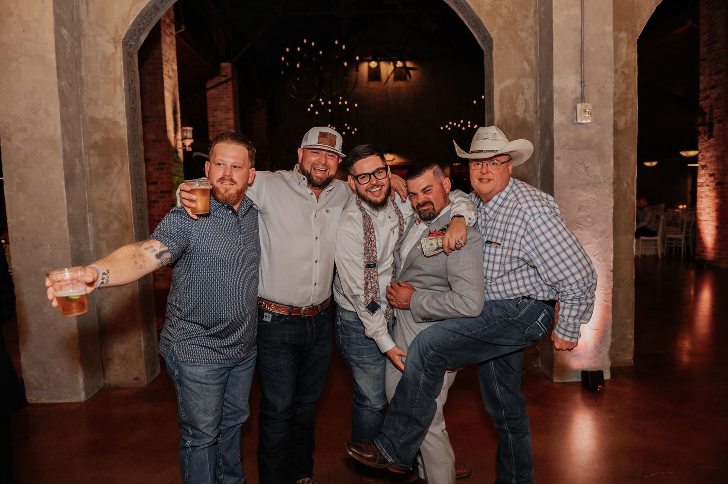 Five men posing and smiling together at a country western wedding reception in a rustic venue.