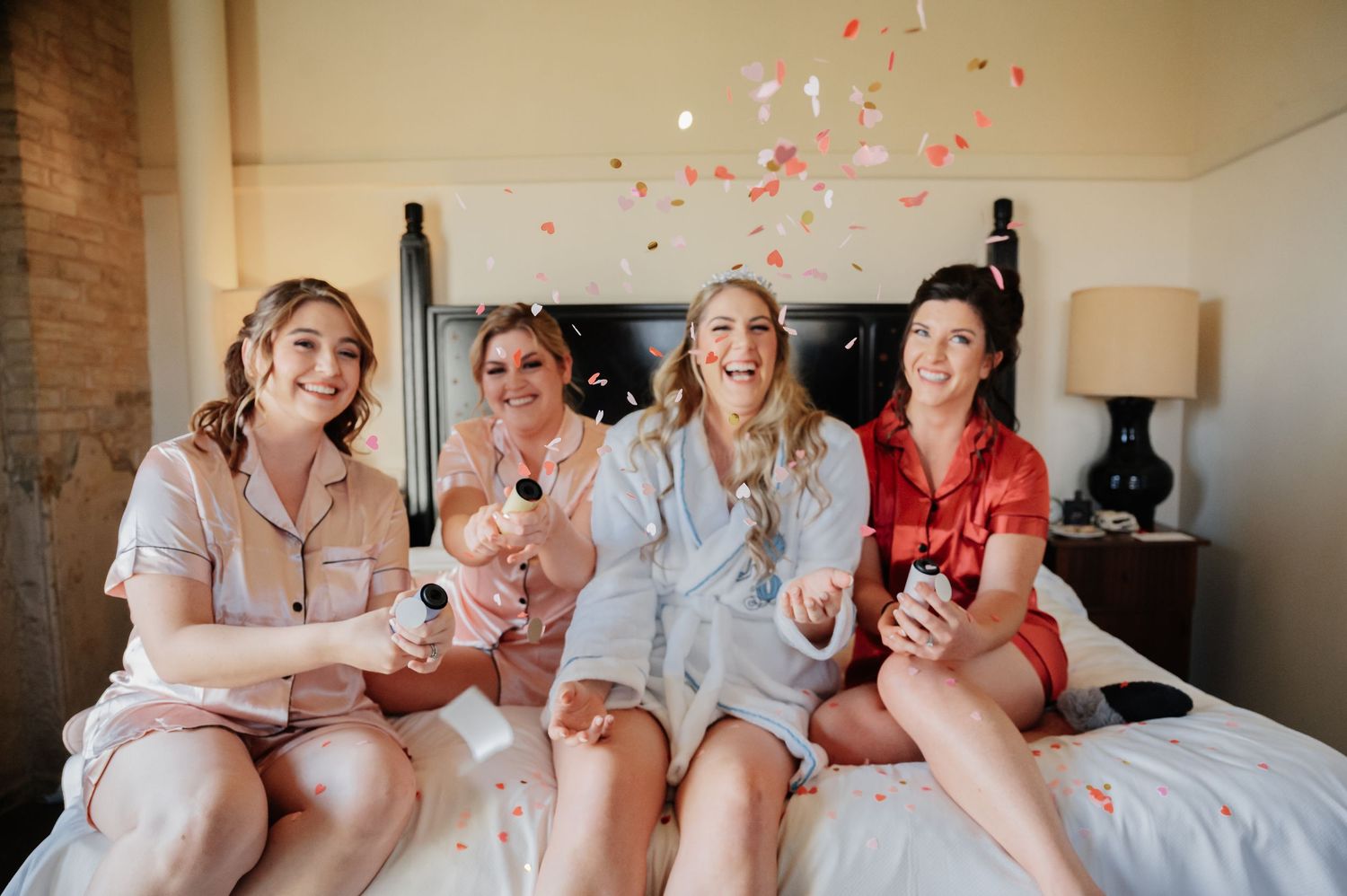 Four bridesmaids in pajamas celebrating on a bed with champagne and confetti during a bridal party getting-ready session.