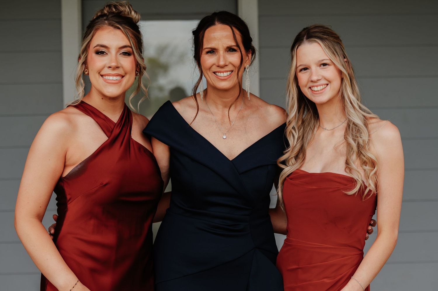 Three women in formal dresses pose together smiling, wearing red and navy outfits at a special occasion.