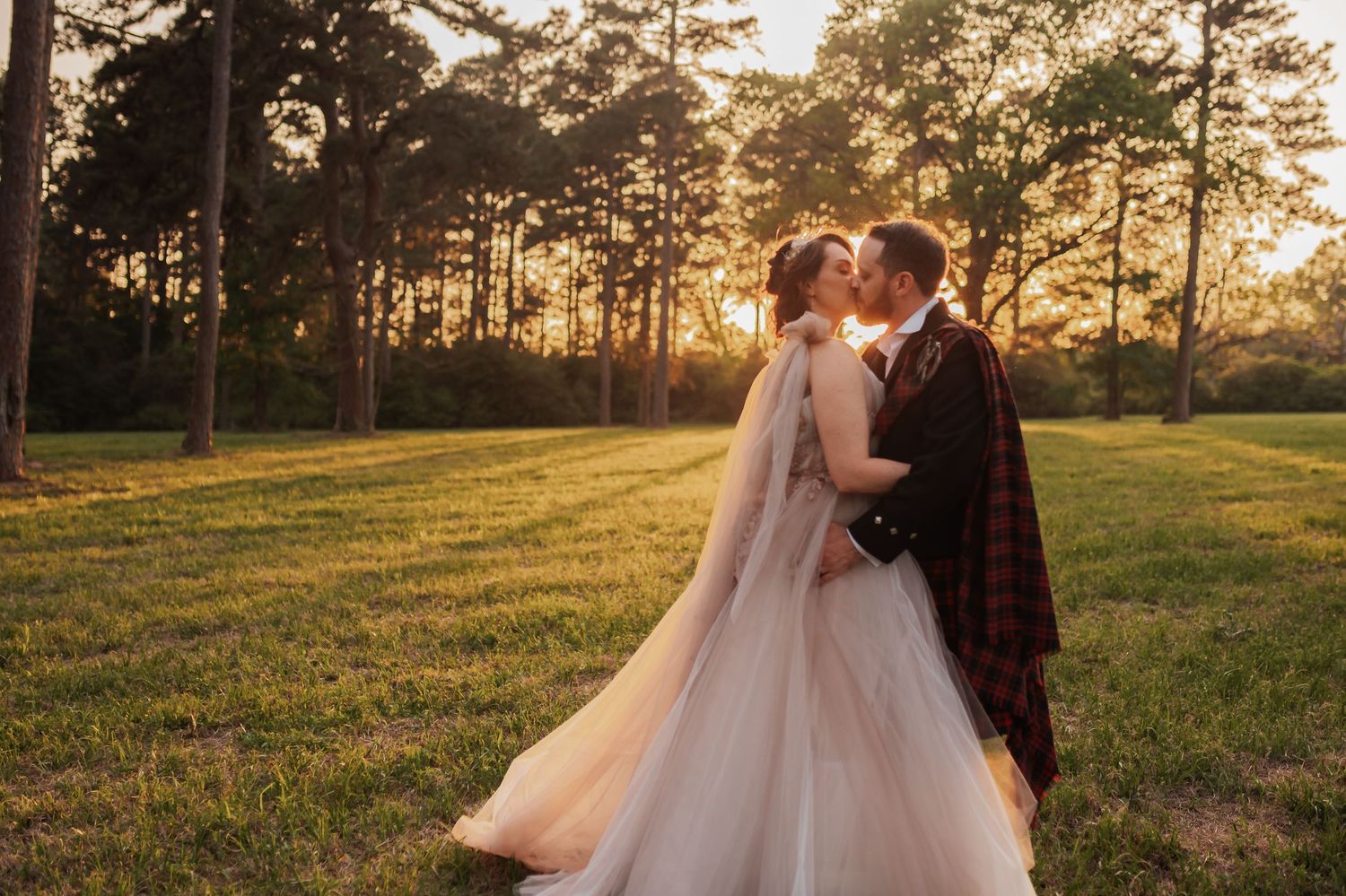 Bride and groom share a romantic sunset kiss in a lush green field, bride in flowing white gown, groom in tartan kilt.