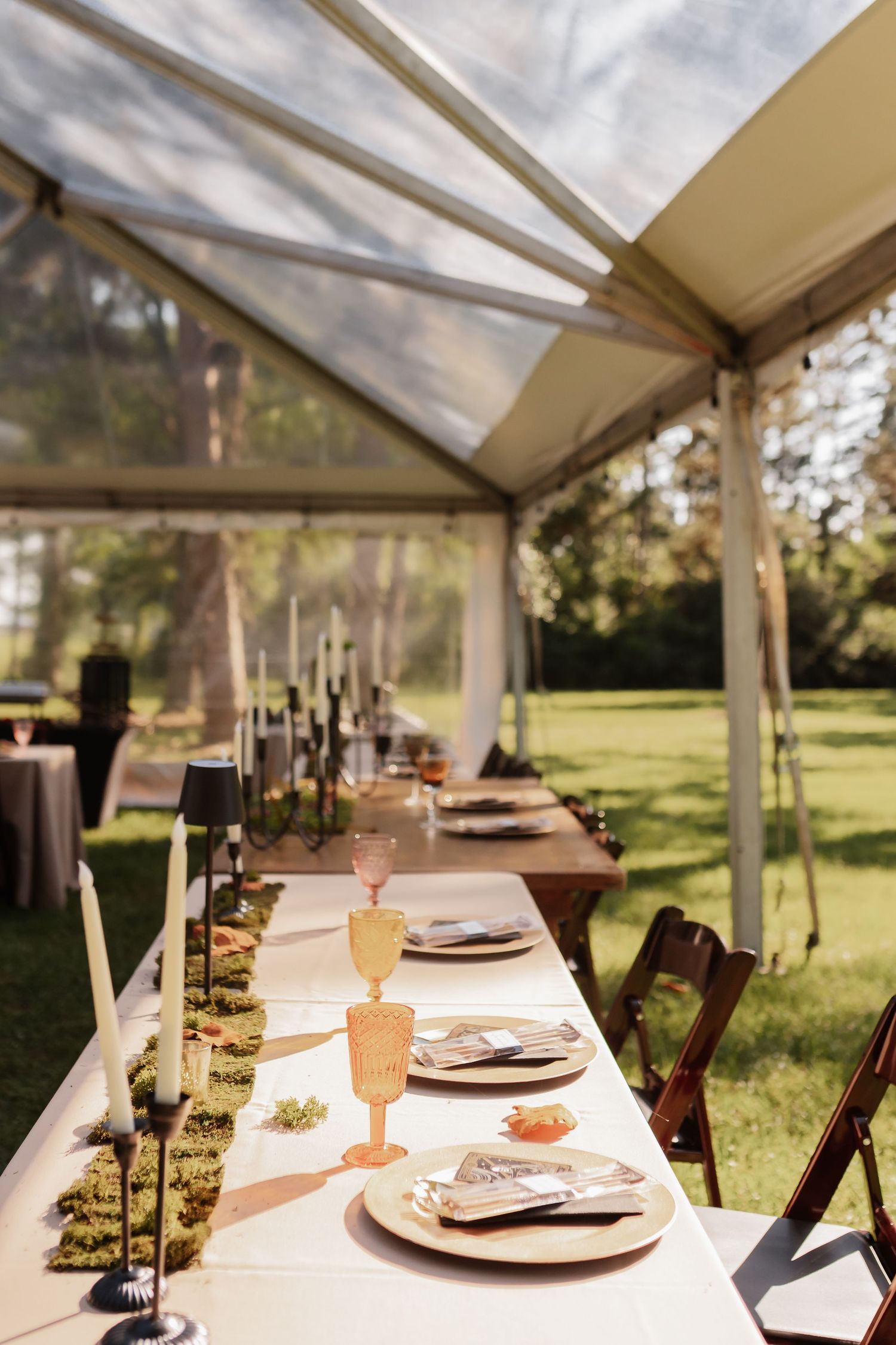 Elegant outdoor wedding reception table set under a clear tent with candles, moss runner, and colored glassware on green lawn.