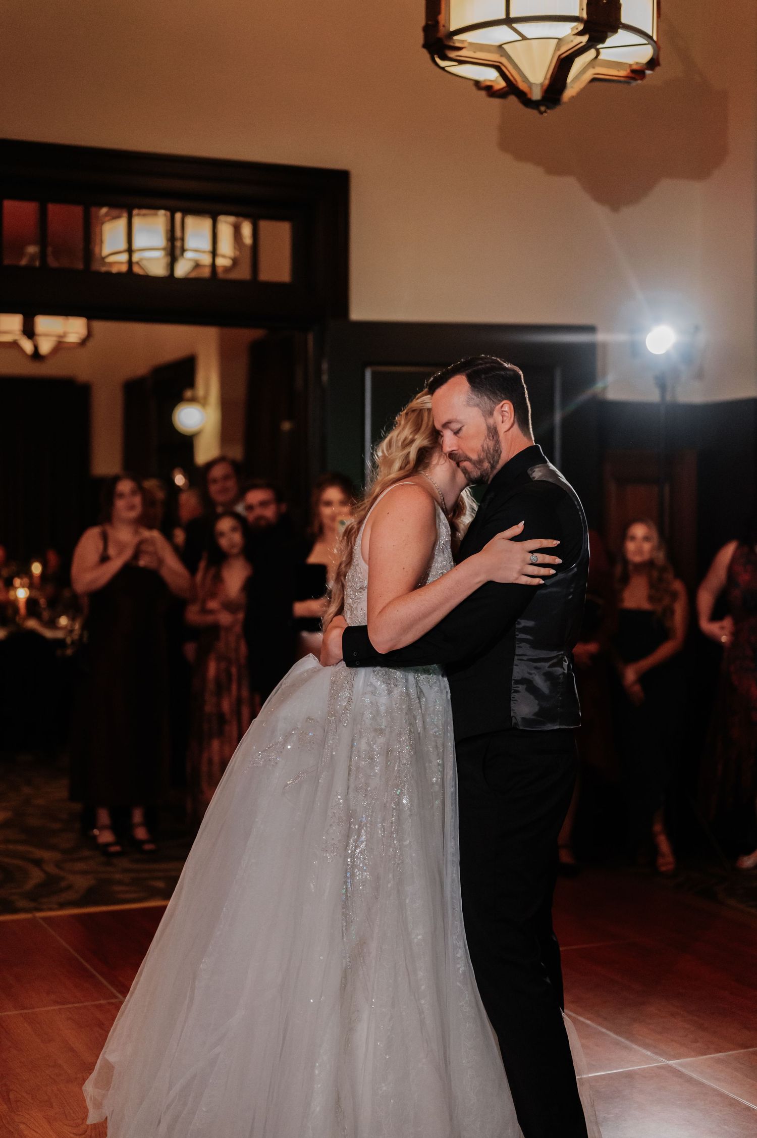 Bride and groom share an intimate first dance at their wedding reception, surrounded by guests in elegant attire.