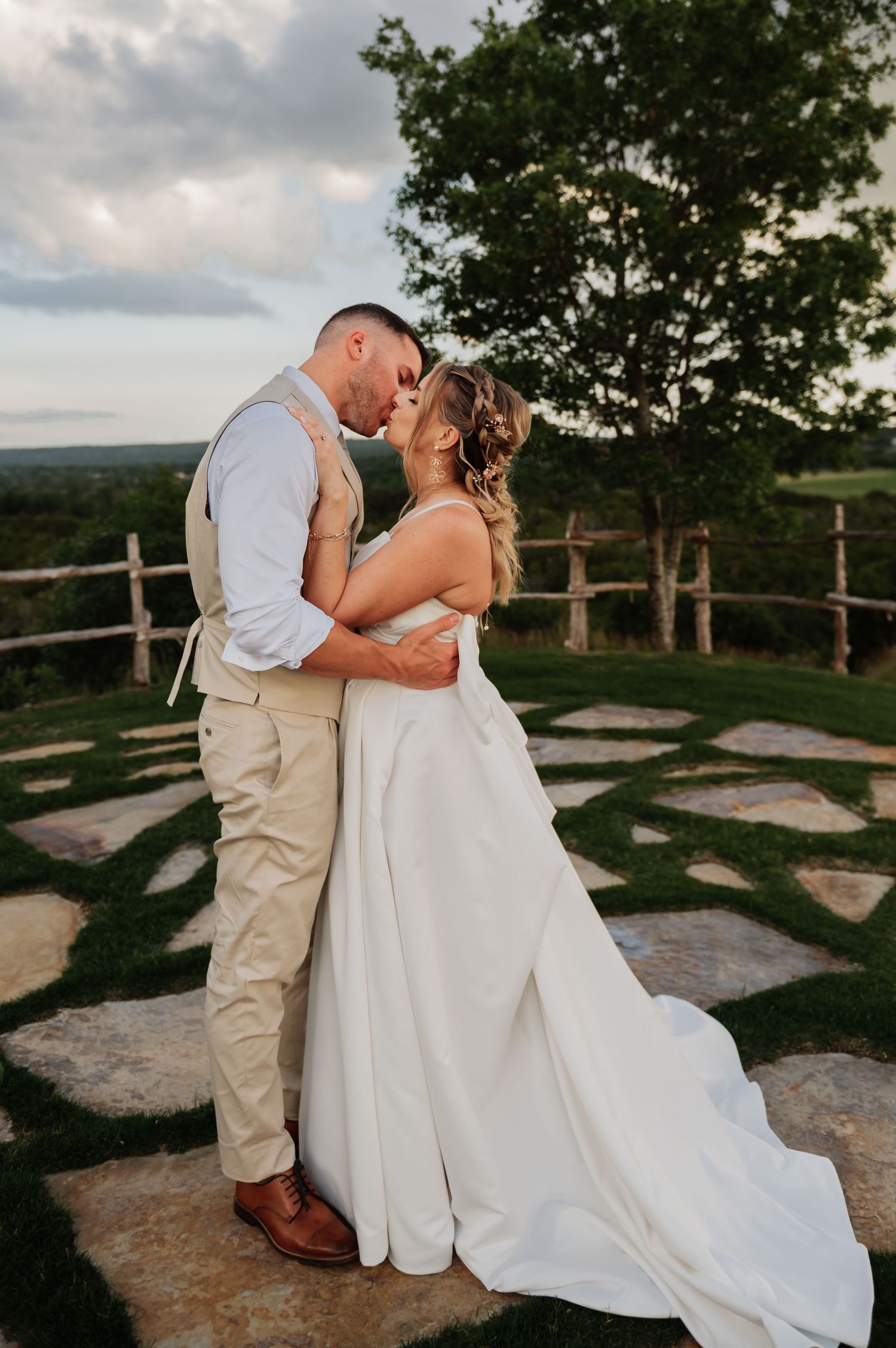 Bride and groom sharing a romantic kiss in white gown and tan suit on stone patio with scenic outdoor backdrop.