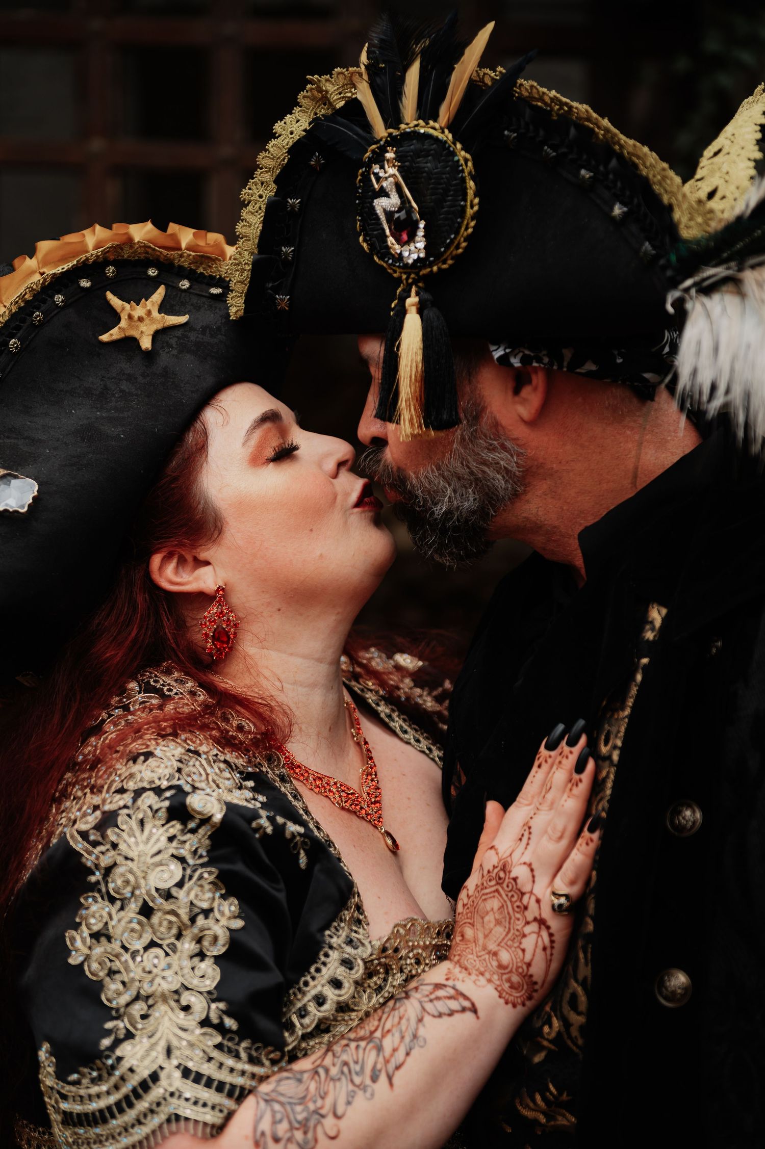 Couple sharing a kiss in ornate pirate attire with gold embroidery and tricorn hats at a themed wedding.