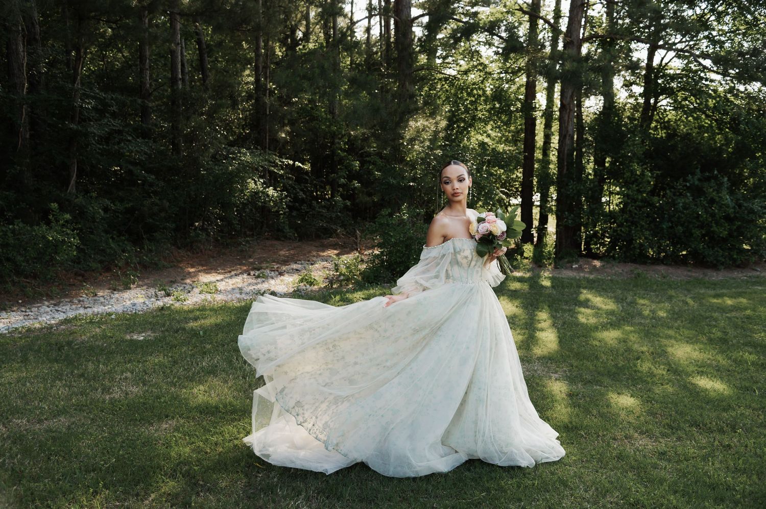 Bride in a flowing white lace gown twirls in a sunlit garden surrounded by lush green trees.