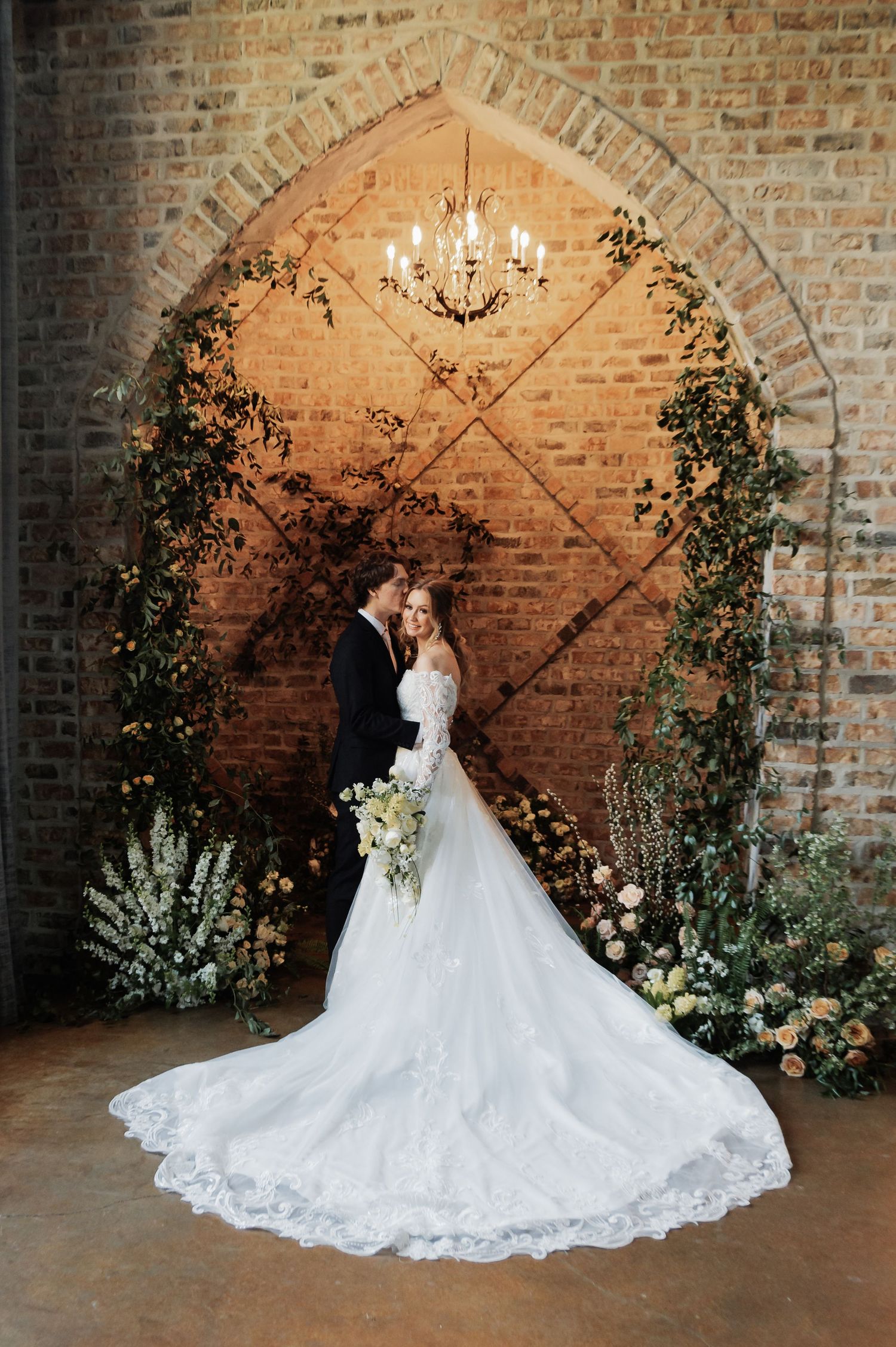 Couple embraces under a floral arch in a rustic brick venue with lush greenery and a chandelier overhead.
