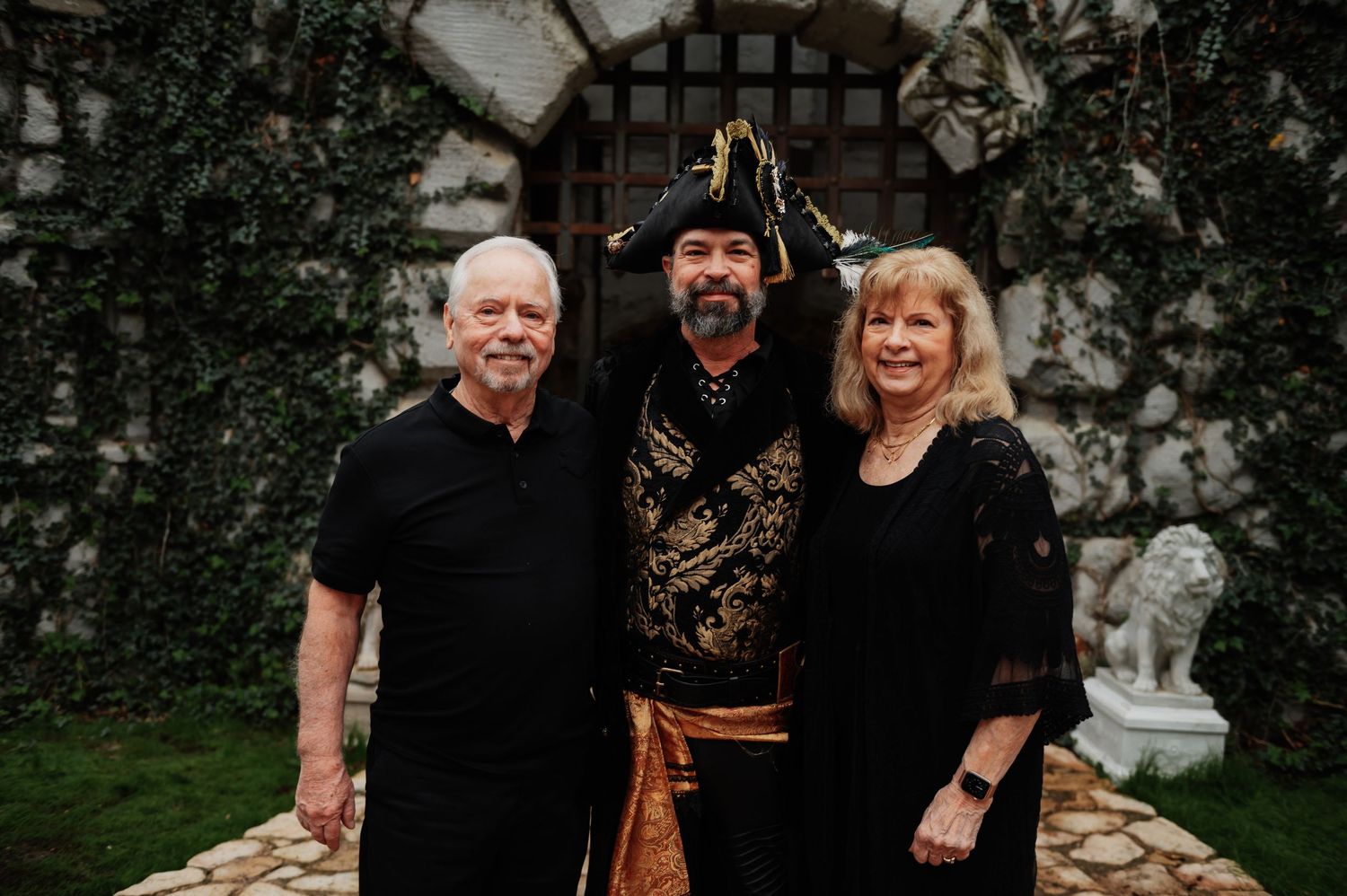 Three adults pose outdoors, one dressed in pirate costume, against a stone wall backdrop.