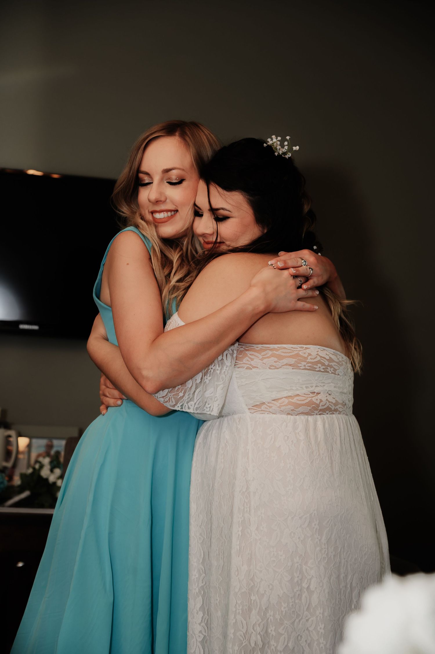 Bride in white lace gown shares emotional hug with bridesmaid in turquoise dress on wedding day.