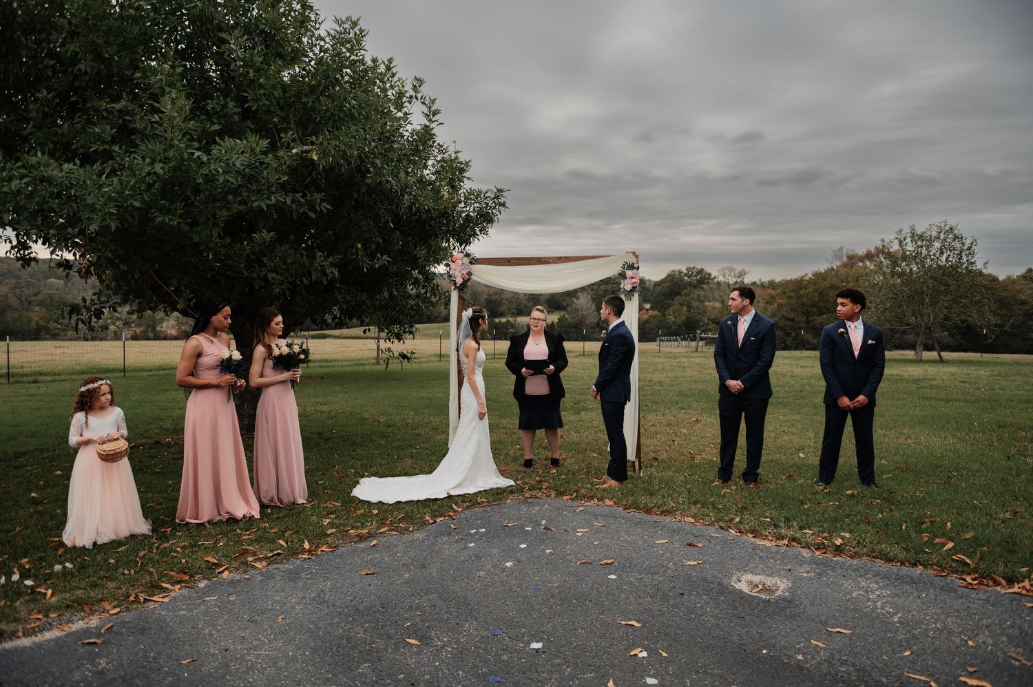 Wedding ceremony outdoors on cloudy day with bride, groom, officiant, bridesmaids in pink, and groomsmen under floral arch.