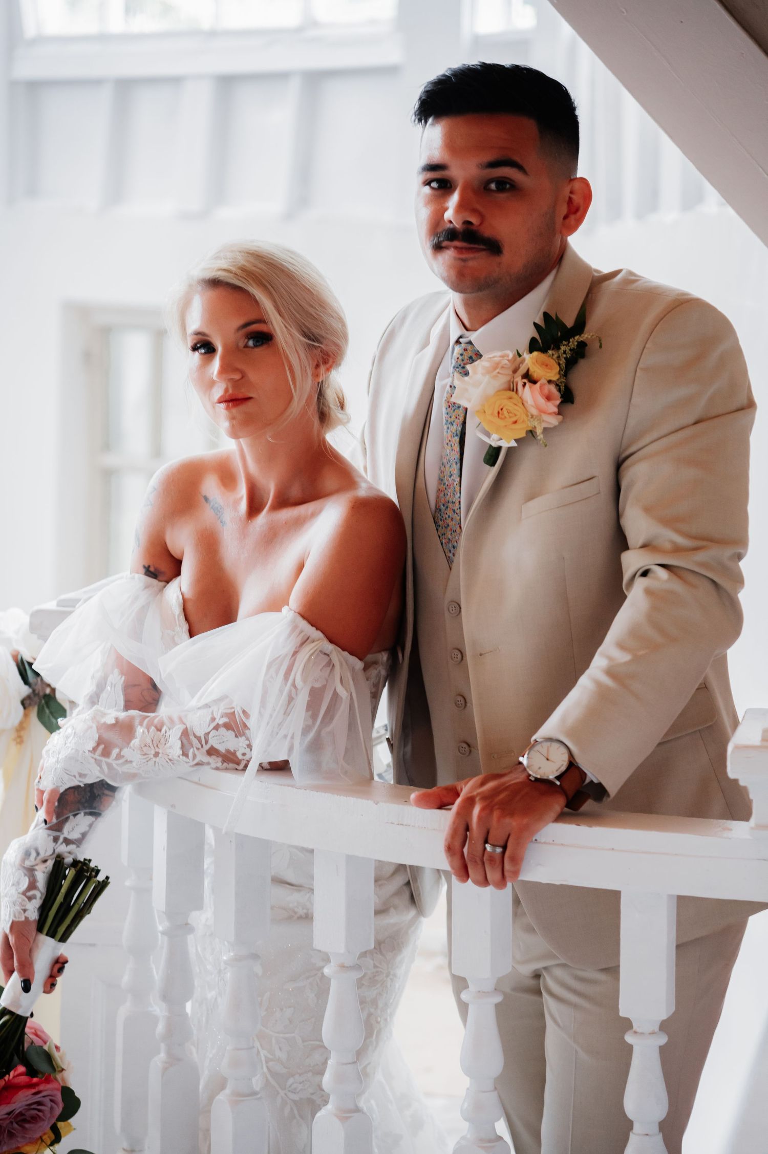 Bride in lace gown and groom in beige suit with floral boutonniere pose elegantly on white staircase railing.
