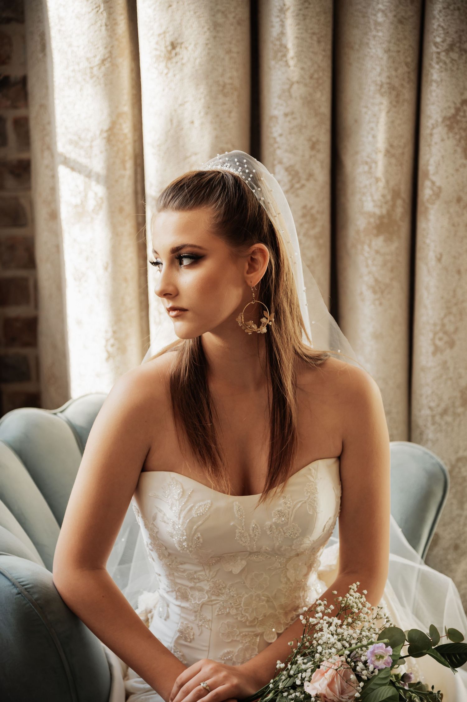 Elegant bride in strapless lace gown with veil and hoop earrings, holding floral bouquet while seated by curtained window.