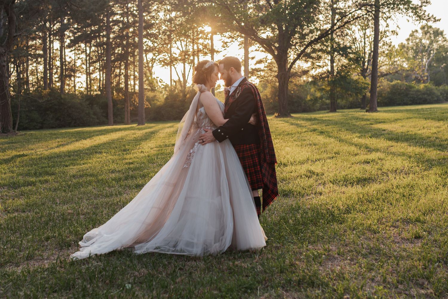 Bride and groom share a romantic moment at golden hour in a lush green park, with sunlight filtering through tall trees.