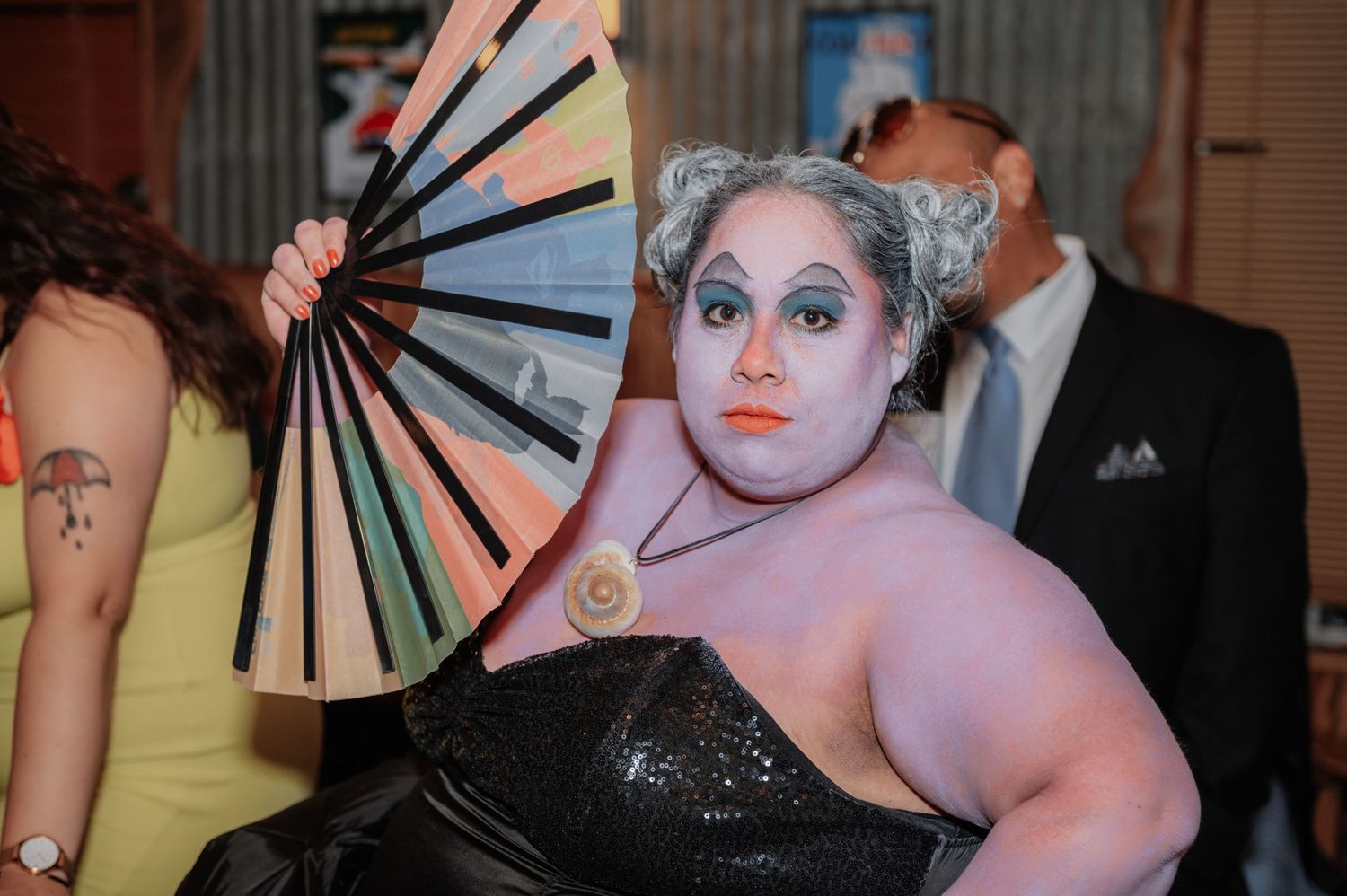Drag performer in black sequin dress holds decorative fan with dramatic makeup at an evening event.