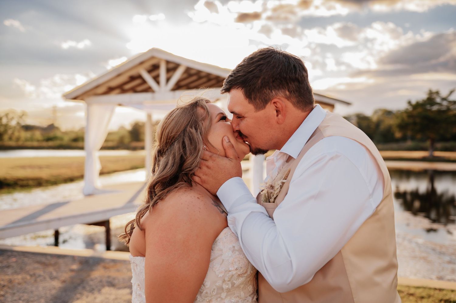 Bride and groom share a romantic kiss near a white pavilion by a lake at sunset on their wedding day.