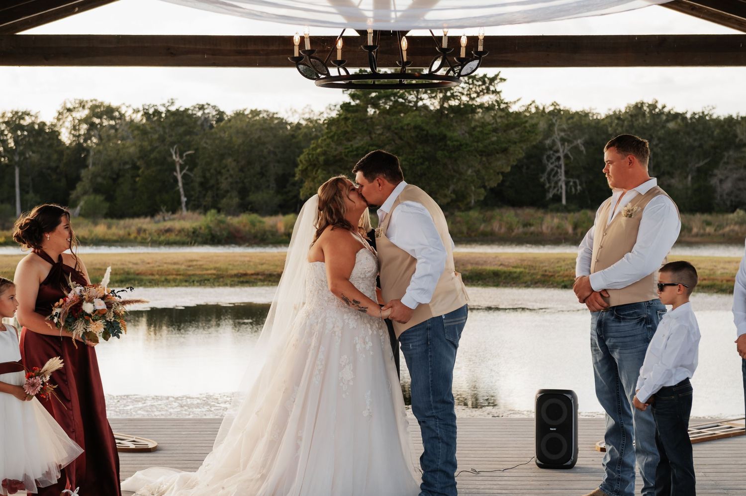 Couple kisses during outdoor lakeside wedding ceremony surrounded by bridal party in burgundy dresses and tan vest.