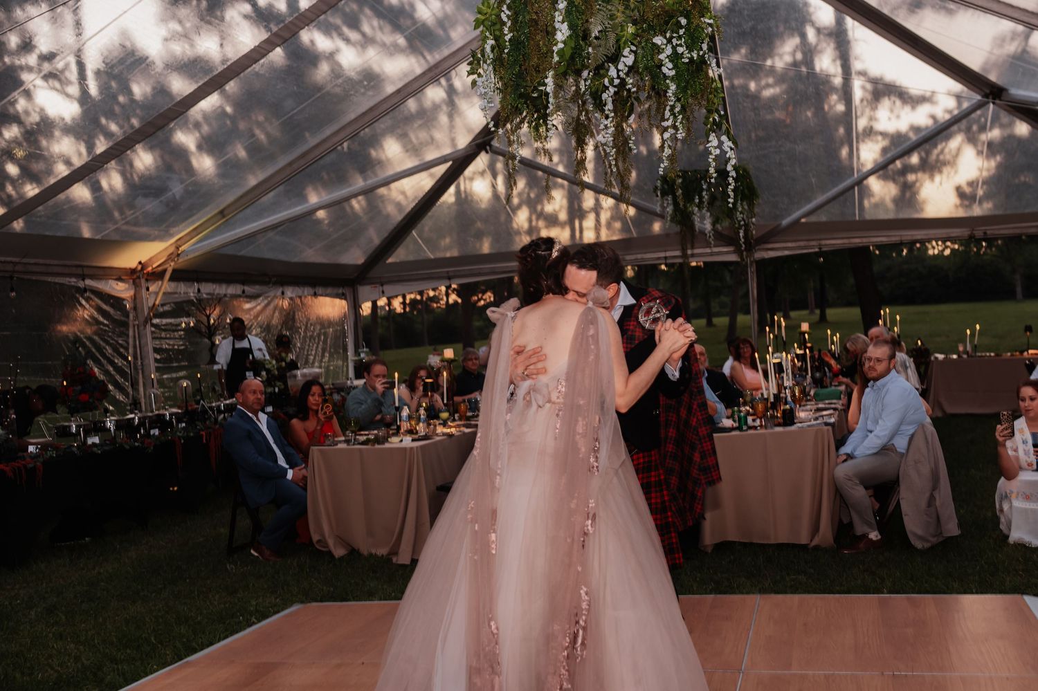 Bride and groom share their first dance under a clear tent with hanging greenery and candlelit tables at their wedding reception.