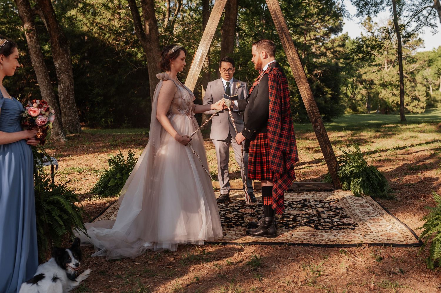 Bride and groom exchange vows outdoors under a wooden arch, groom wearing a red tartan kilt, officiant standing behind them.