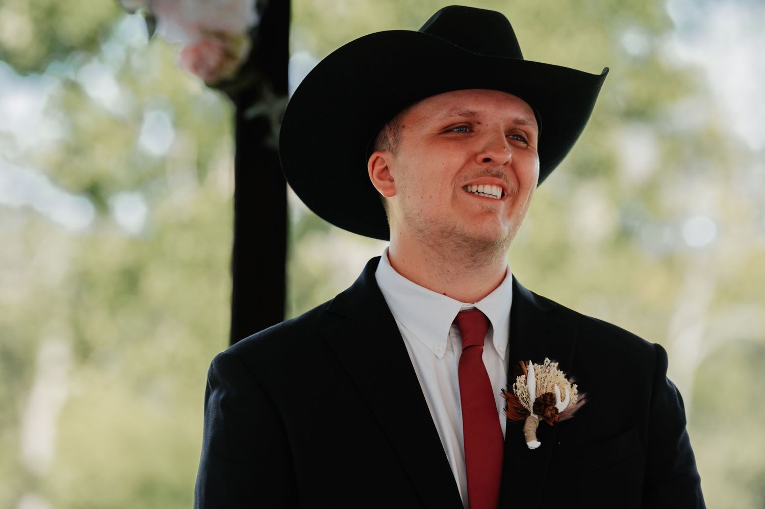 Man in black cowboy hat and suit with red tie and boutonniere smiling at outdoor wedding ceremony.