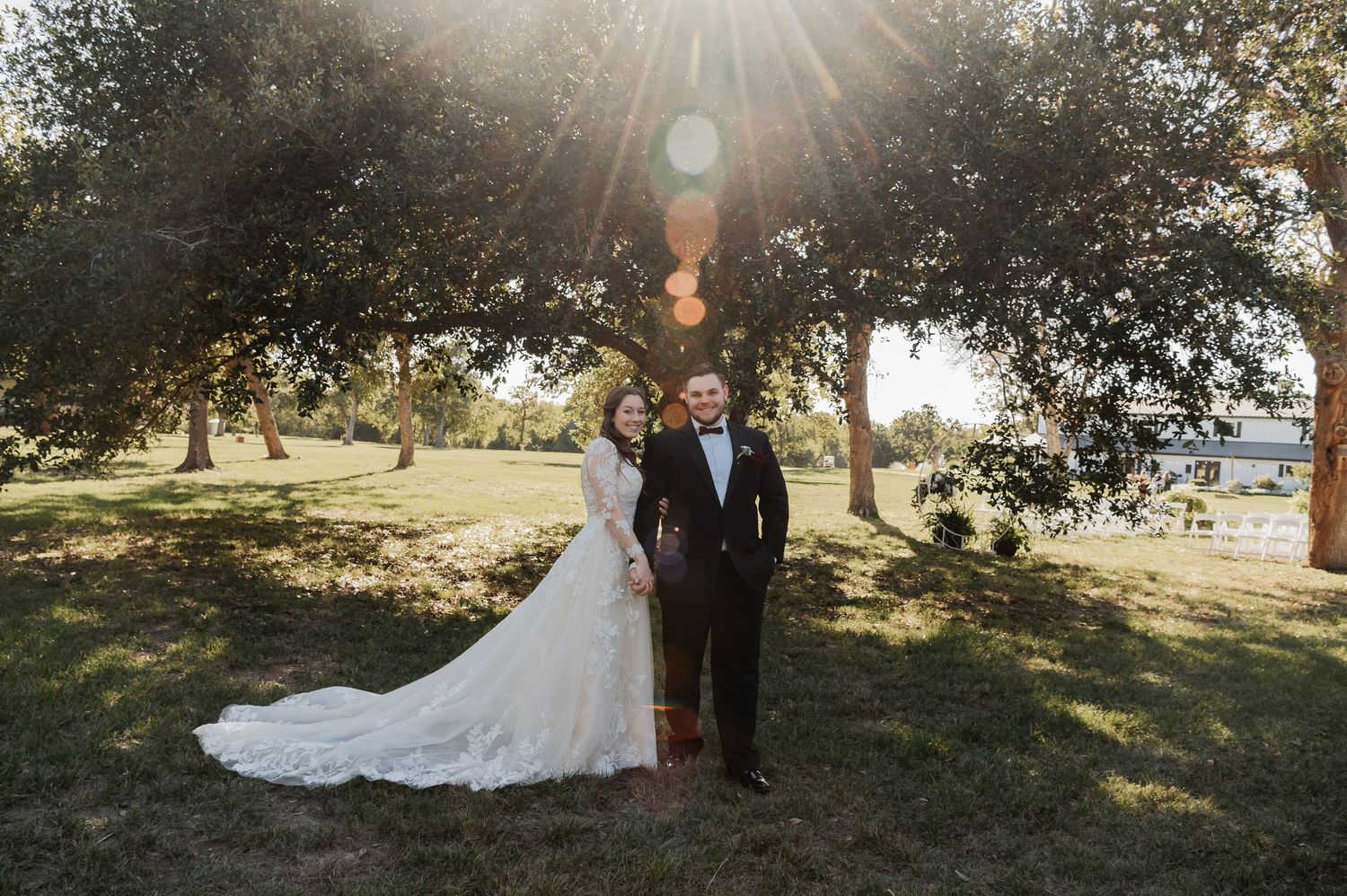 Bride and groom standing under sunlit trees on lush green grass, golden hour sunbeams shining through the canopy.