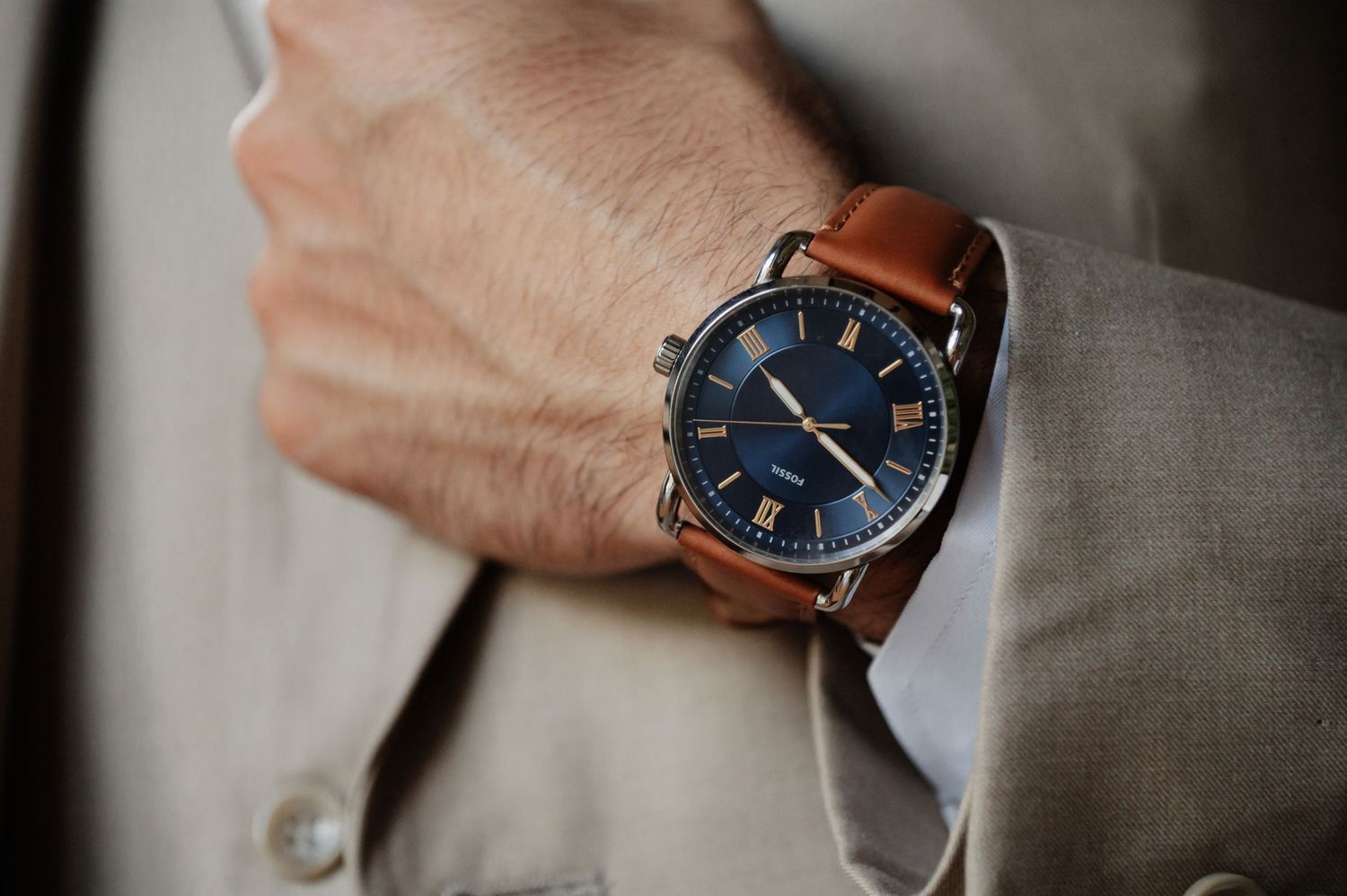 Man wearing a classic blue-dial watch with brown leather strap and silver case on his wrist over a beige suit.