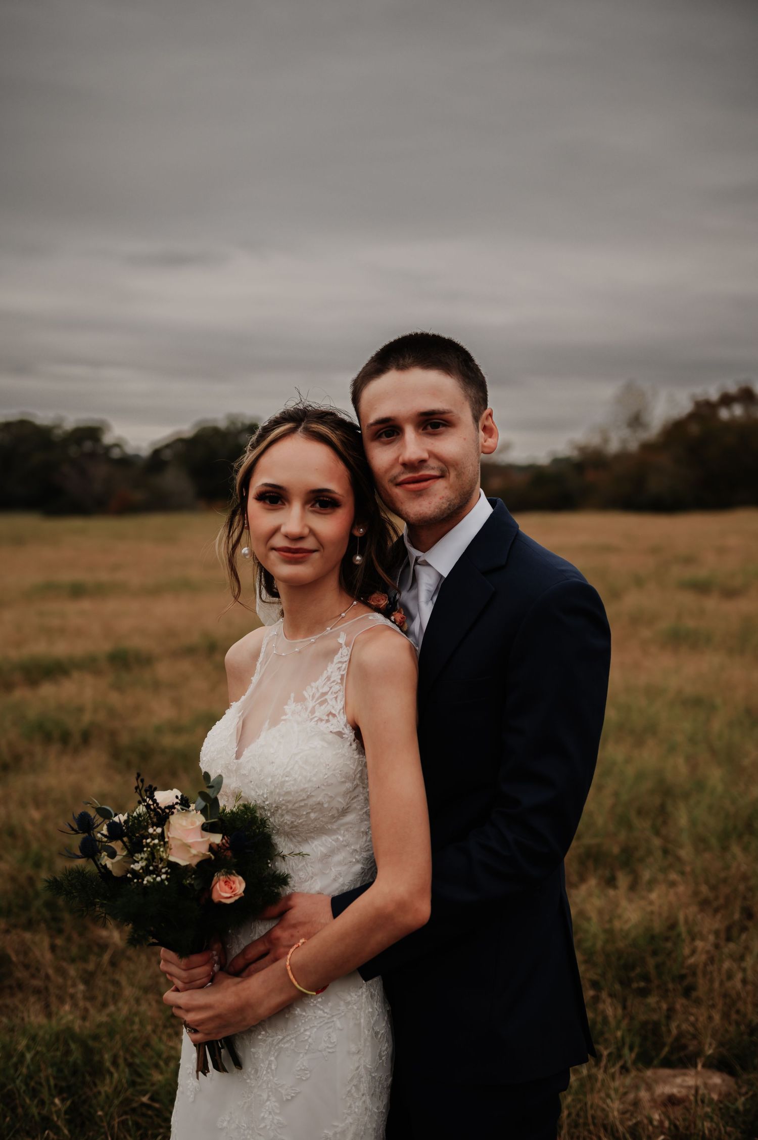 Bride and groom posing in a golden field on their wedding day, bride holding a bouquet of pink and white flowers.