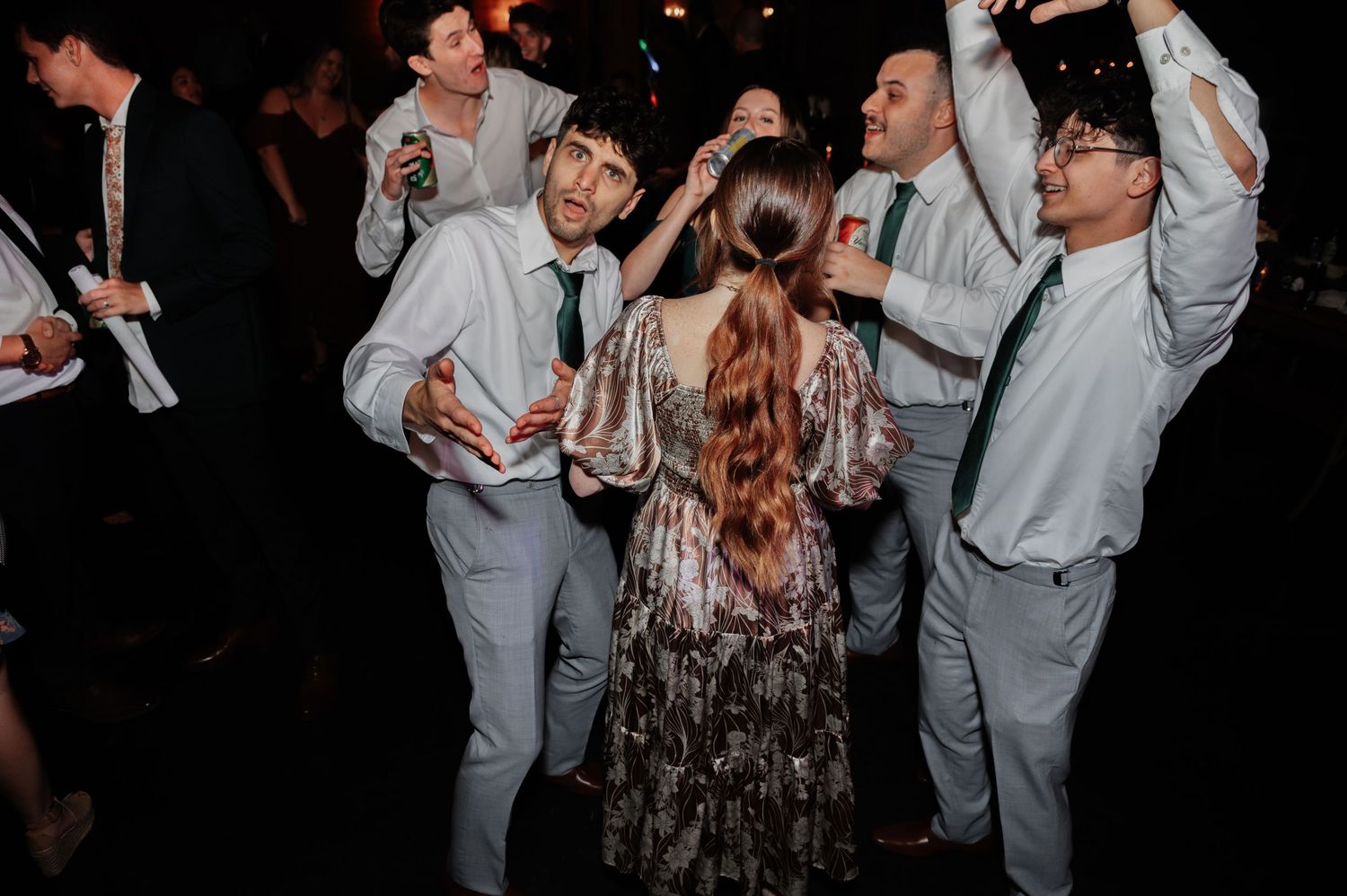 Wedding guests in light suits and green ties dancing joyfully with a woman in a sequined dress at a reception.