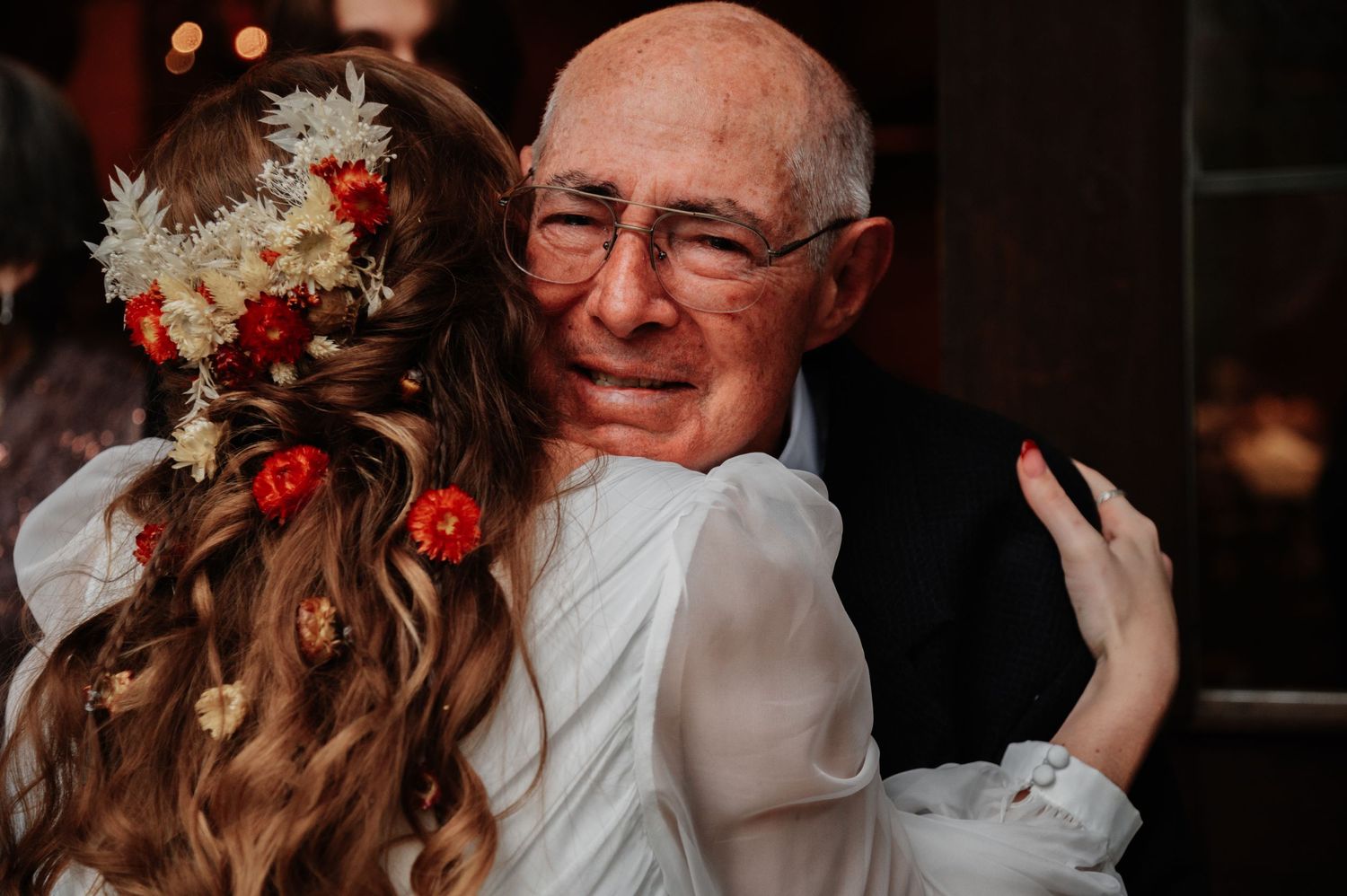 Bride with floral hair accessories shares an emotional hug with elderly man at wedding reception.