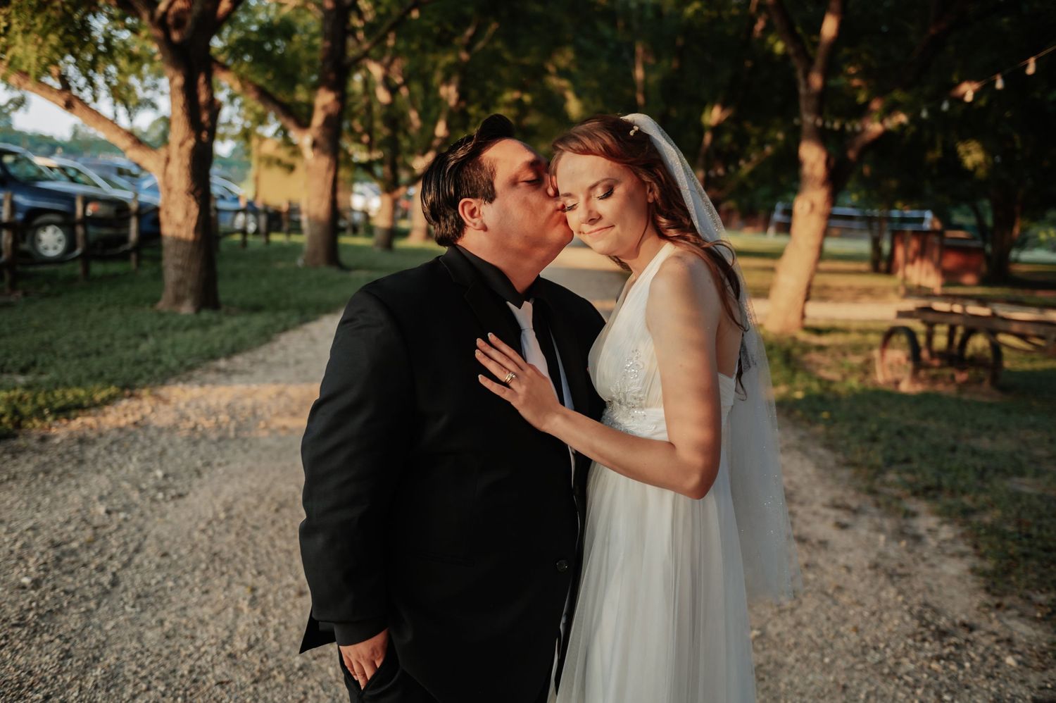 A bride and groom share a tender kiss outdoors at their wedding venue surrounded by trees on a sunny day.
