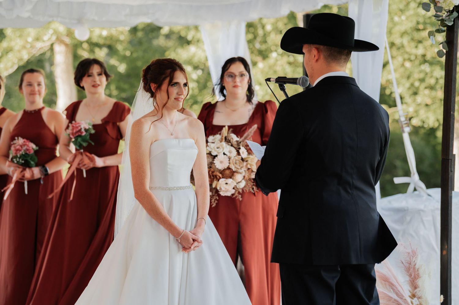 Bride in white gown holds bouquet at outdoor wedding ceremony with bridesmaids in rust dresses.